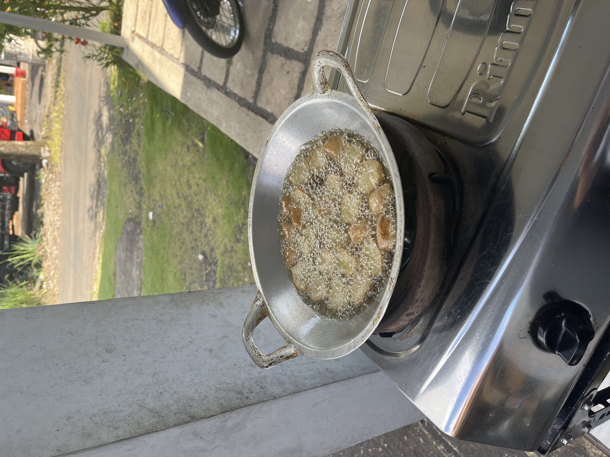 In Jambearum, Indonesia, a close-up shot shows food being fried in a wok on a gas stove. The wok, made of aluminum, sits atop a rusty burner. The food, appearing to be small, golden-brown pieces, is submerged in bubbling oil, creating a lively surface. The gas stove is stainless steel, with a black knob visible on the front. In the background, a paved area with grass and a road is visible, with a red truck and some trees suggesting an outdoor setting.