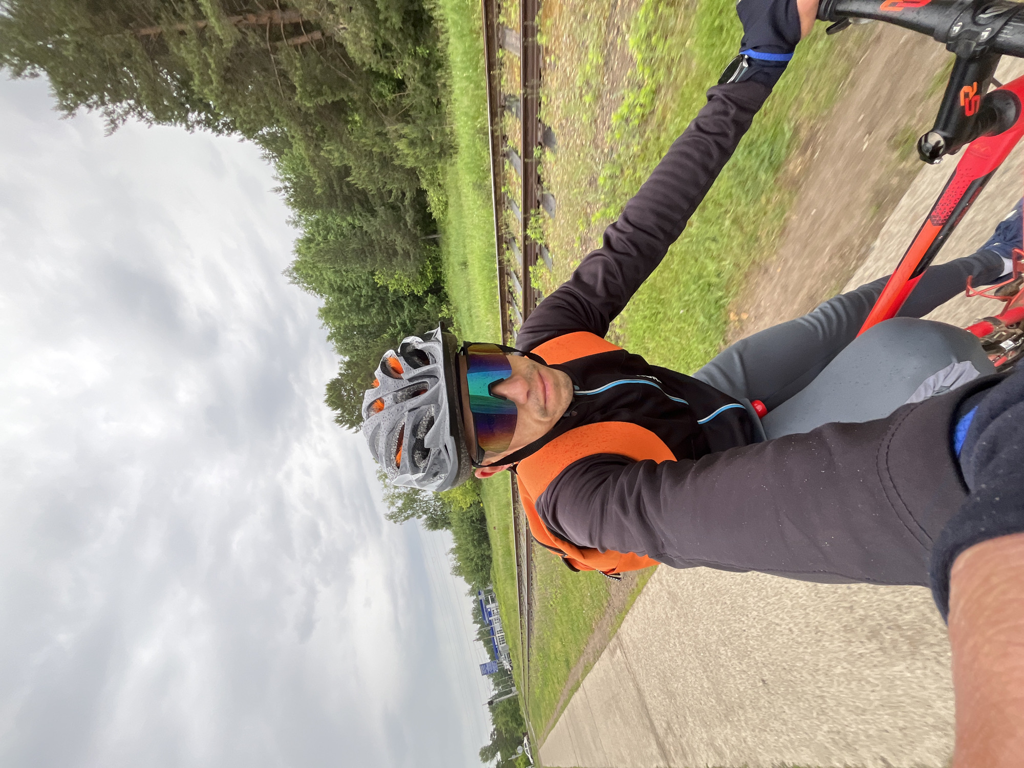 A male cyclist captures a first-person selfie while riding a red and black mountain bike in Chaikovsky, Russia. The cyclist is seen from the chest up, wearing a grey and orange helmet with visible water droplets, colorful mirrored cycling glasses, a black long-sleeved cycling jersey, and a bright orange sleeveless vest or jacket. He has a neutral to slightly smiling expression.
The scene unfolds alongside a paved path or road, which is visible in the foreground. Adjacent to the path, a strip of green grass leads to a set of railway tracks that run parallel into the distance. Beyond the tracks, a dense line of coniferous trees forms the background. A small blue and white building, possibly a station or utility structure, is visible further along the tracks. The sky is overcast with light grey clouds, indicating a cloudy daytime. The droplets on the helmet and clothing suggest damp weather, possibly from recent rain. Motion blur in the background indicates the cyclist is moving. Text on the bicycle frame appears to be a stylized "2S" or "2F".