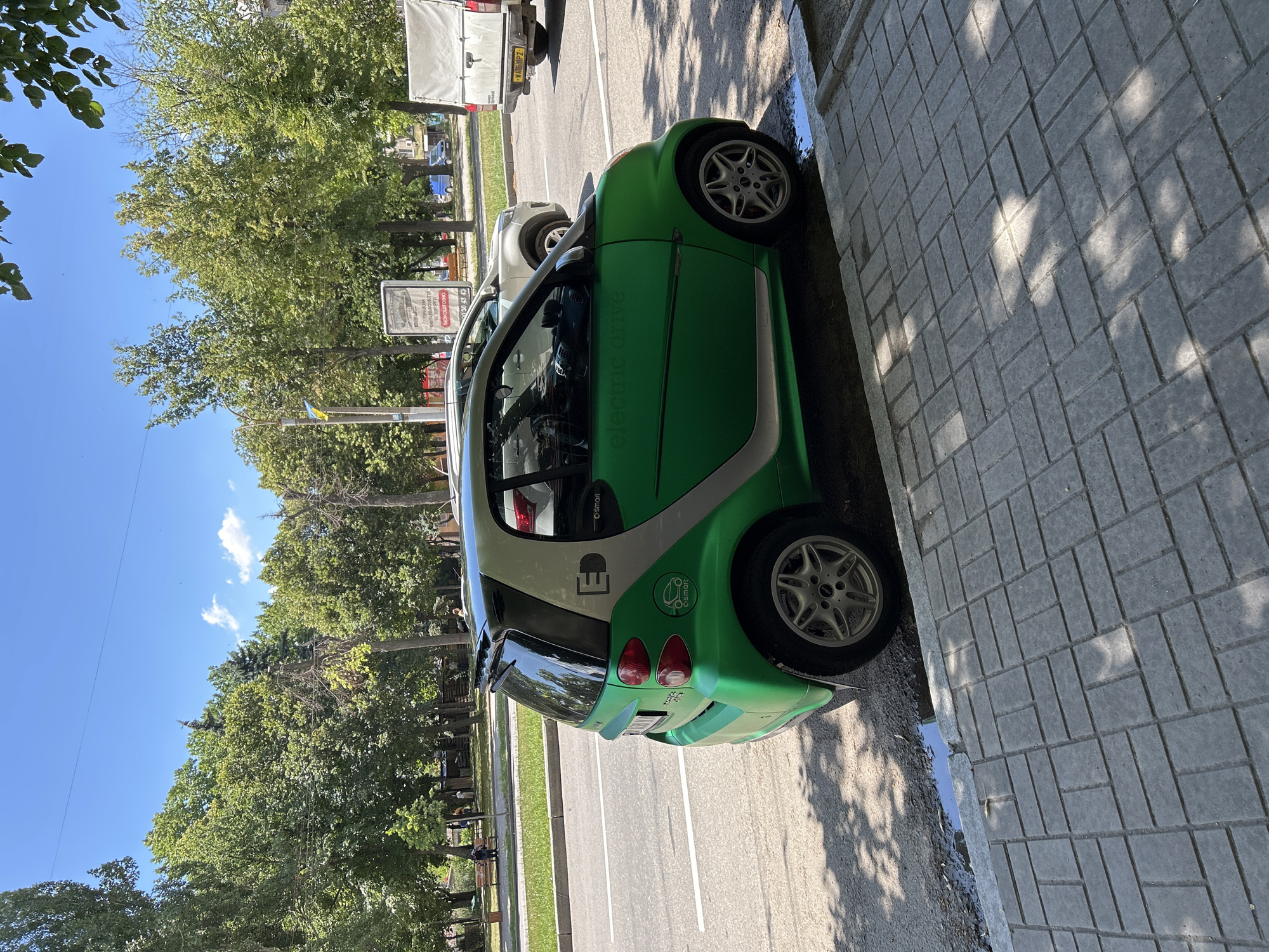 A green Smart electric car is parked on a sidewalk next to a street in Zaporizhzhia, Ukraine. The car has the "e-smart" logo on its side and a stylized "E" on the rear pillar. The sidewalk is paved with grey rectangular bricks, and there are shadows of trees on it. The street has white lane markings, and other vehicles are visible in the background, including a white SUV and a white van. Trees line the street, and there is a park-like area with benches and people in the background. The sky is blue with a few white clouds.