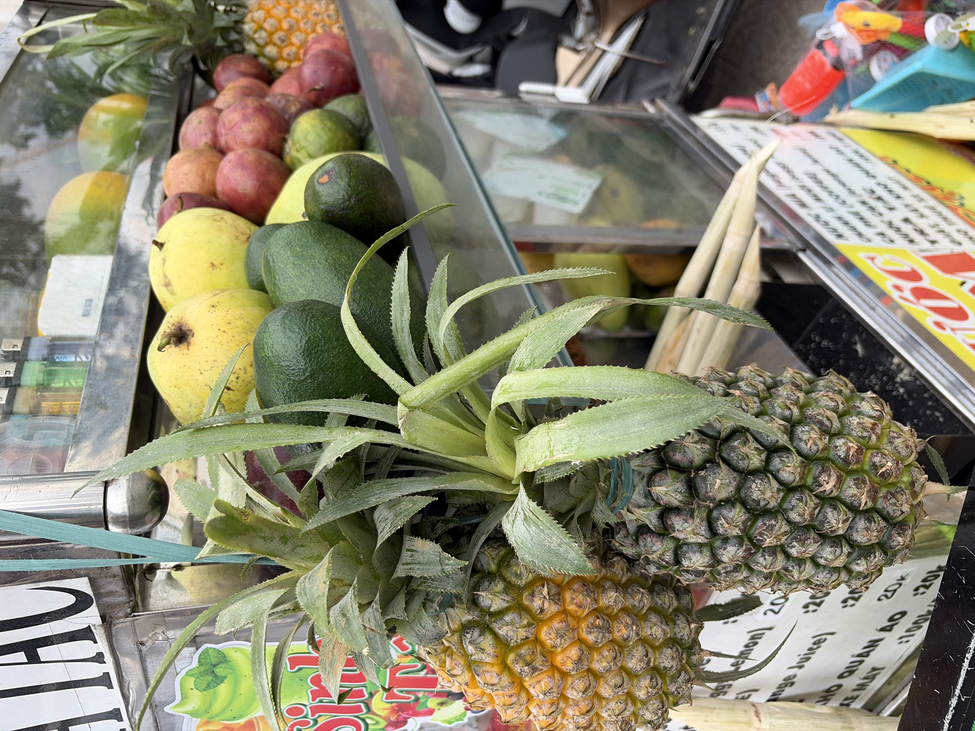 A street food cart is loaded with a variety of fruits, including pineapples, avocados, guavas, and passion fruits. The cart displays a menu in Vietnamese, with prices listed in Vietnamese Dong (k). The background reveals the busy street scene of Da Nang, Vietnam, with a hint of traffic and urban activity. The cart appears to be well-maintained and stocked with fresh produce, indicating a thriving business operation. The vibrant colors of the fruits and the lively atmosphere of the street create a sense of a typical day in this bustling Vietnamese city.