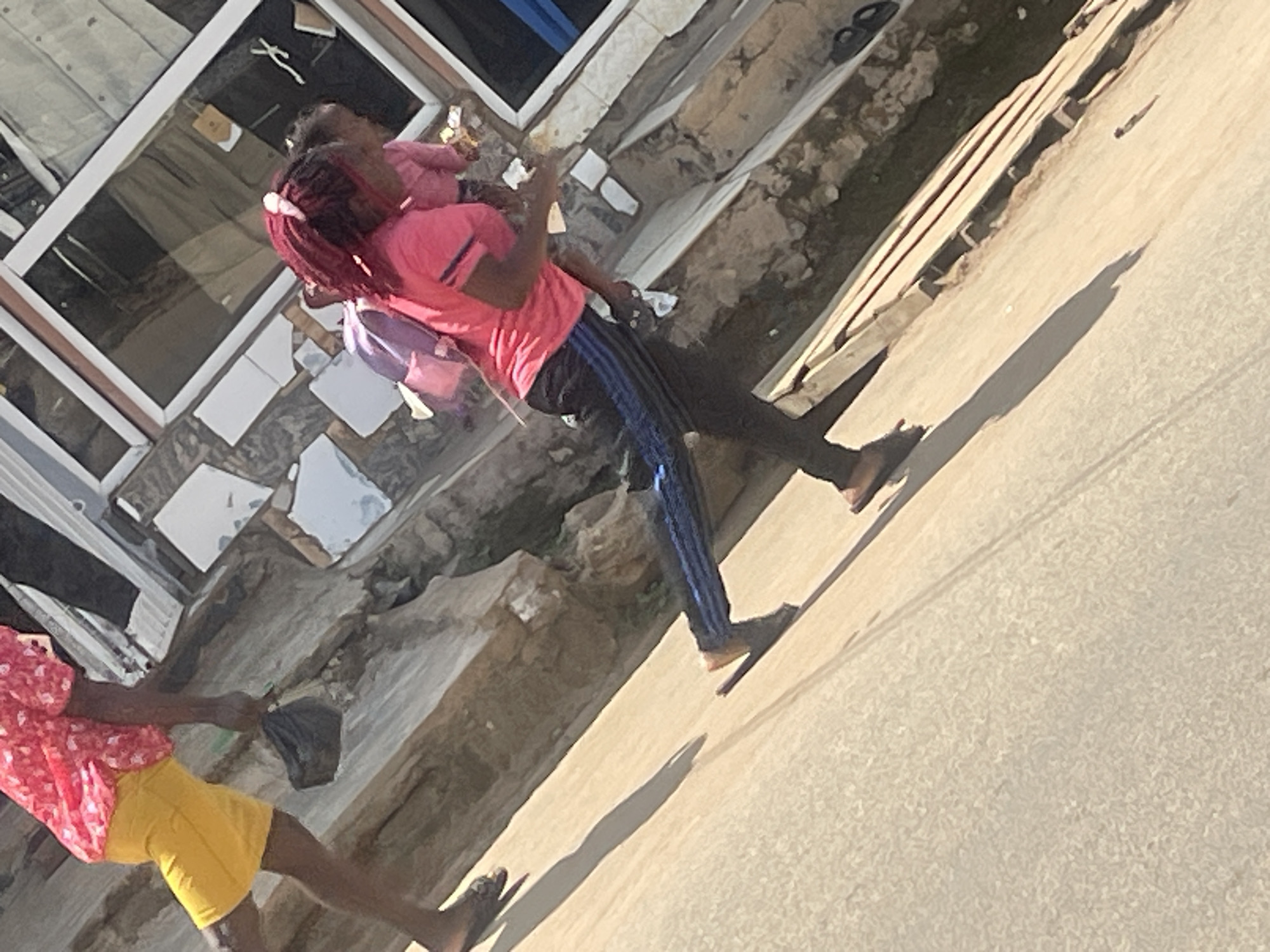 The image shows a woman walking on a street in Abuja, Nigeria. She is wearing a pink shirt and black pants with blue stripes. She has a backpack on her back and is holding a bag in her hand. The woman appears to be walking towards the right of the frame. The street is paved and there are buildings in the background. The lighting suggests it is daytime.