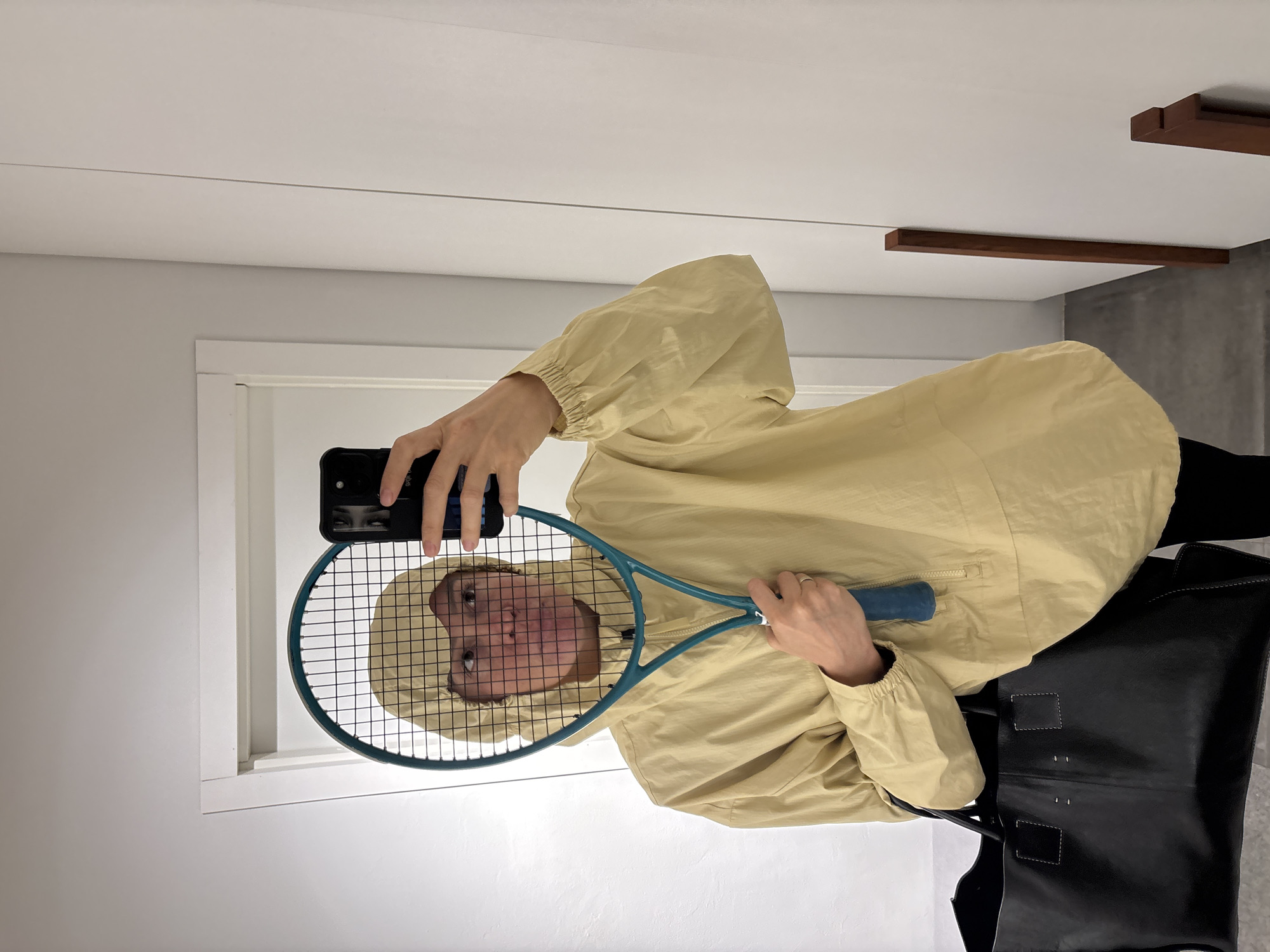A person is holding a tennis racket in front of their face while taking a selfie with a smartphone. The person is wearing a light yellow hooded jacket and a large black tote bag is visible on the right side of the frame. The location appears to be indoors, possibly in a clothing store fitting room, characterized by white walls and a modern, minimalist aesthetic. No information indicates that the image was taken in Chaikovsky, Russia.