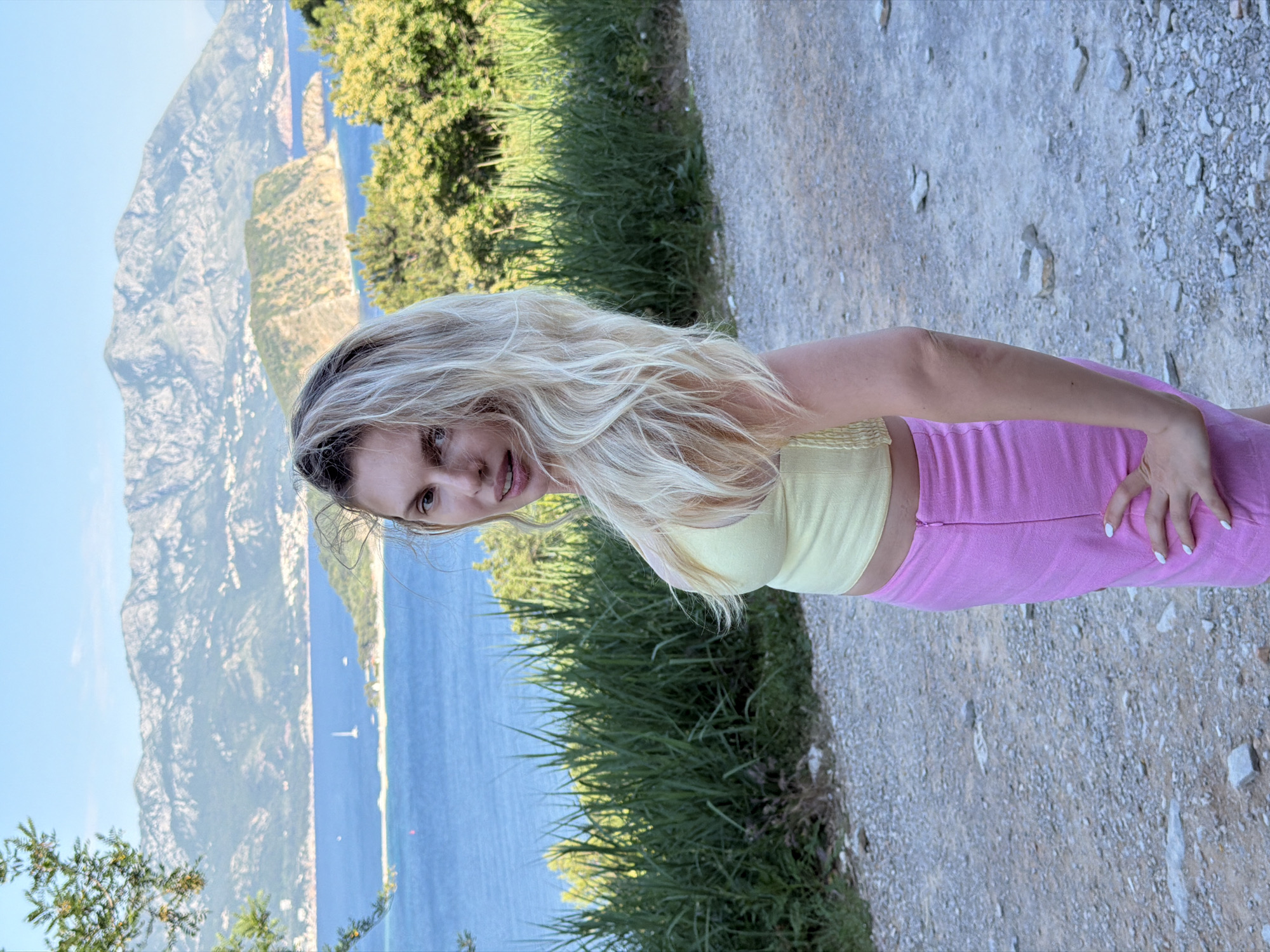 A woman is standing on a gravel path, with a light-green tank top and pink trousers. In the background, there is a body of water with mountains behind it, likely in the city of Budva, Montenegro. The mountains are partially obscured by trees, and the woman is looking directly at the camera.