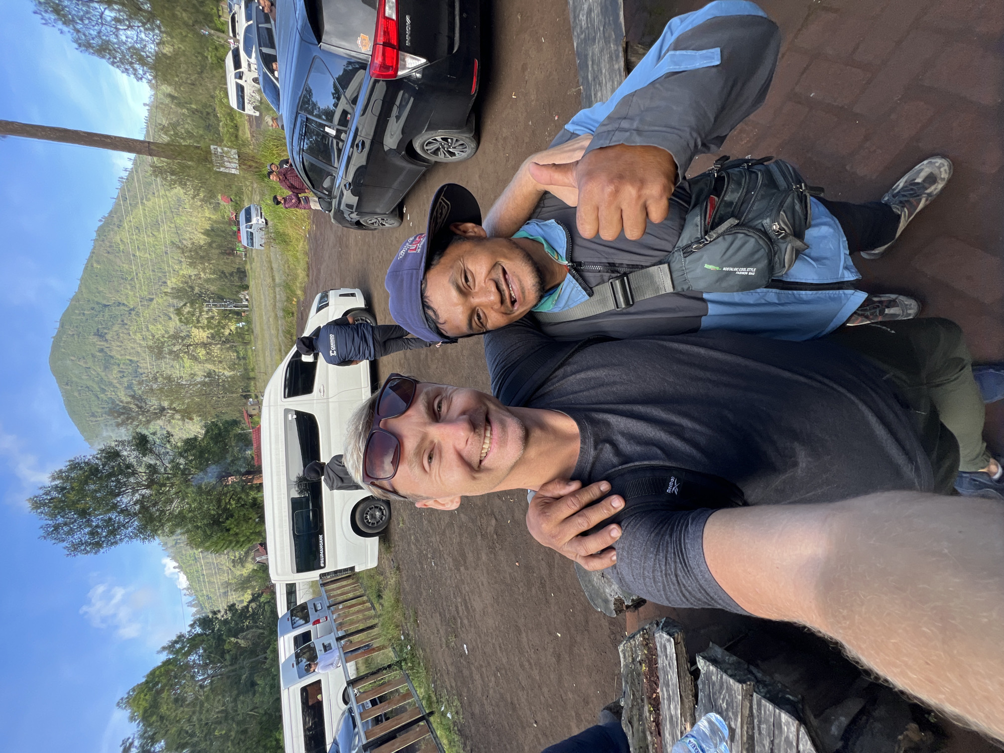 Two men pose for a selfie in an outdoor setting. The man in the foreground wears sunglasses and a dark shirt. The man behind him wears a cap, a blue jacket, and a small bag around his waist; he gives a thumbs-up gesture. The background shows vehicles, including minivans, parked on a dirt surface. A hill or mountain is visible in the distance, partially obscured by trees and foliage. The vans have '#KEGUNUNGANCAK' printed on them. The location is Unknown, Indonesia.