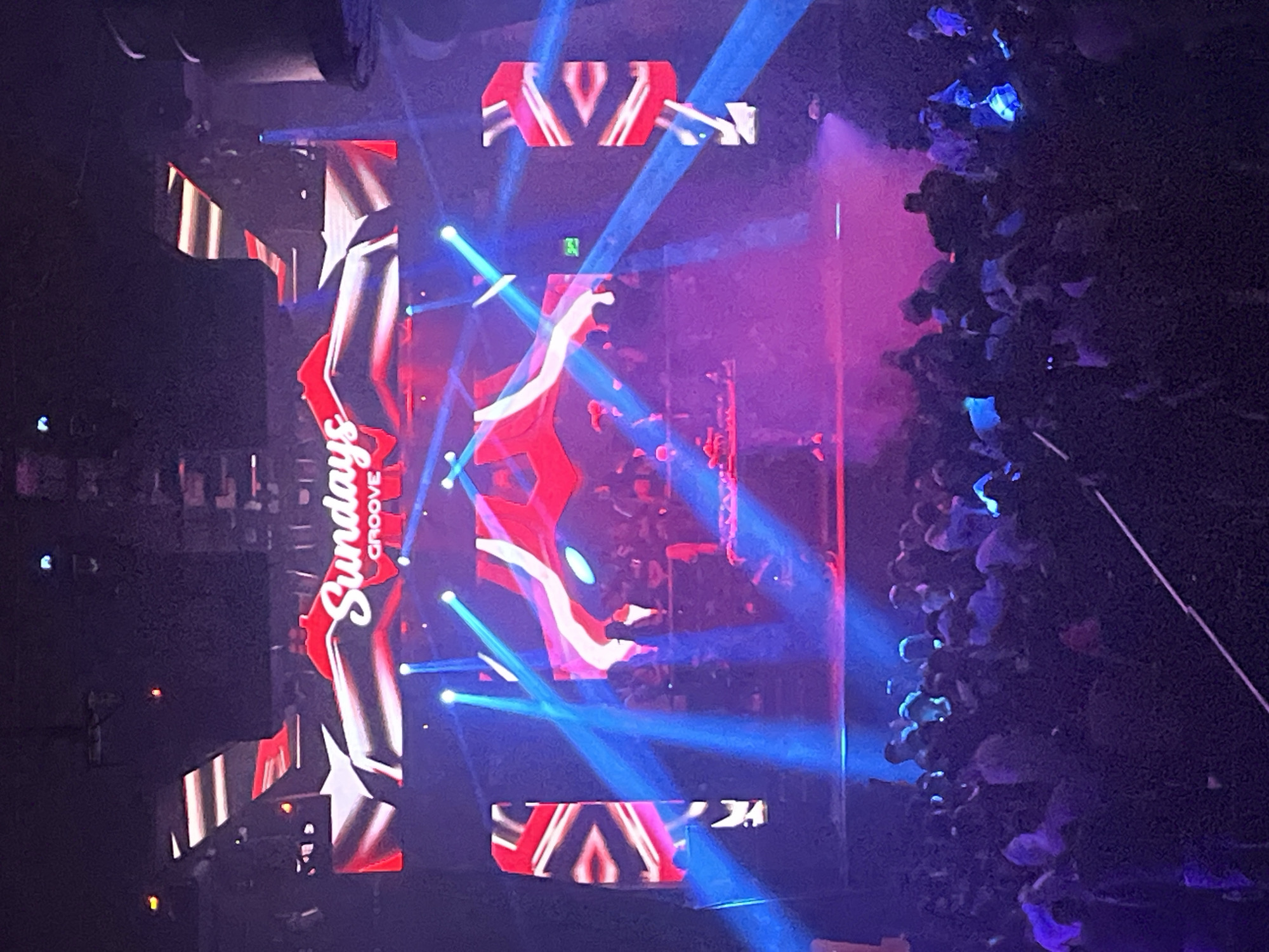 The image depicts a night scene at a club or event likely located in Buenos Aires, Argentina. The environment is dark with strong stage lighting consisting of blue laser beams and pinkish hues from a stage show. There is a large crowd of people, densely packed, primarily visible on the right side of the image. They are lit by stage lighting.
A vertically oriented screen displays "Sundays Groove" in a stylized font. Several decorative screens with a red and white V-shaped pattern are present. The lighting suggests an energetic, party atmosphere.