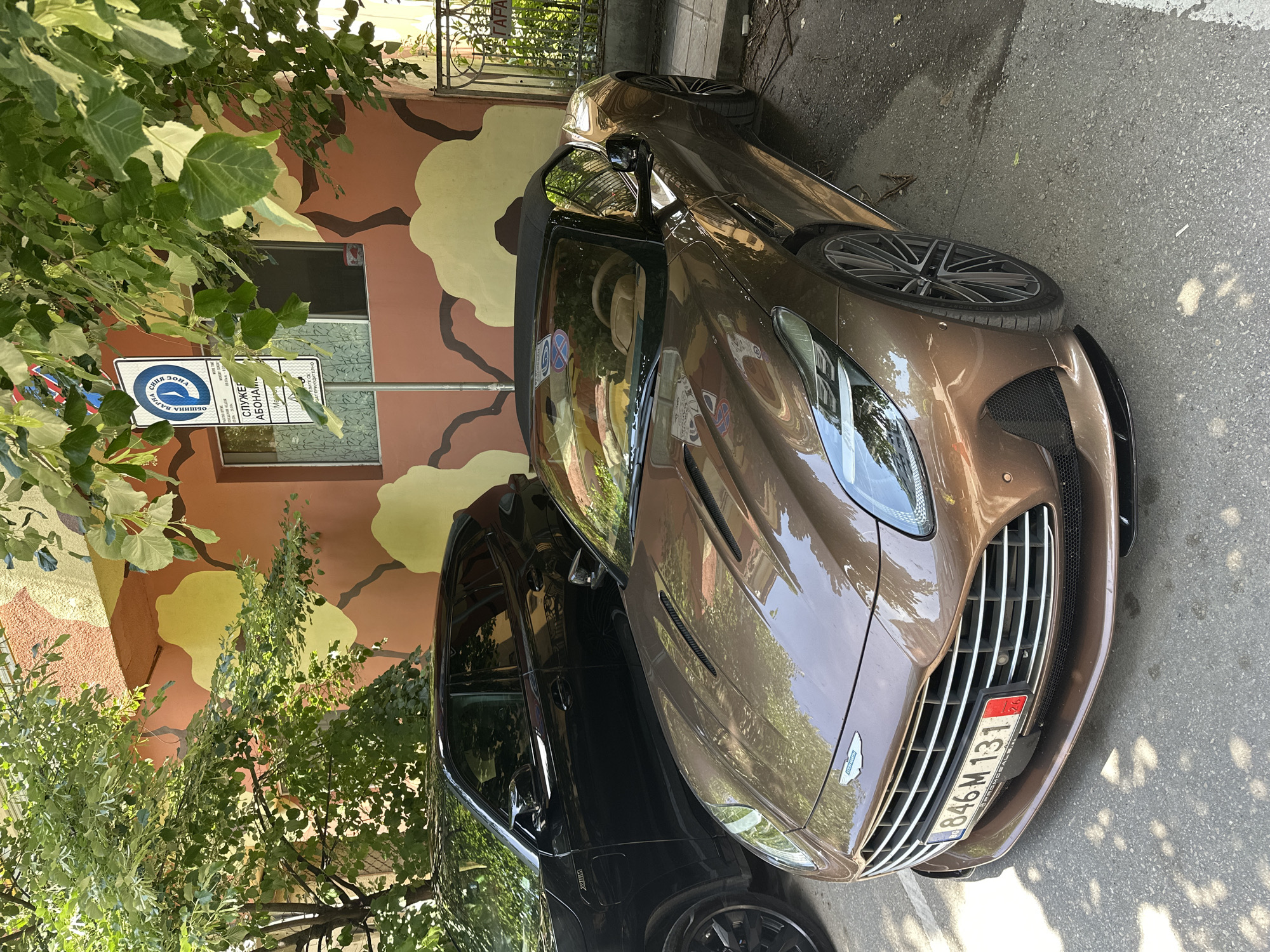 A bronze-colored Aston Martin DBX is parked on a street in Varna, Bulgaria. The license plate reads "В 846 М 131" with a Bulgarian flag and "BG" emblem. A black SUV is parked behind it. A sign for a paid parking zone is visible behind the car. The background features a building facade with an unusual camouflage-like painting. Trees are partially obscuring the view.