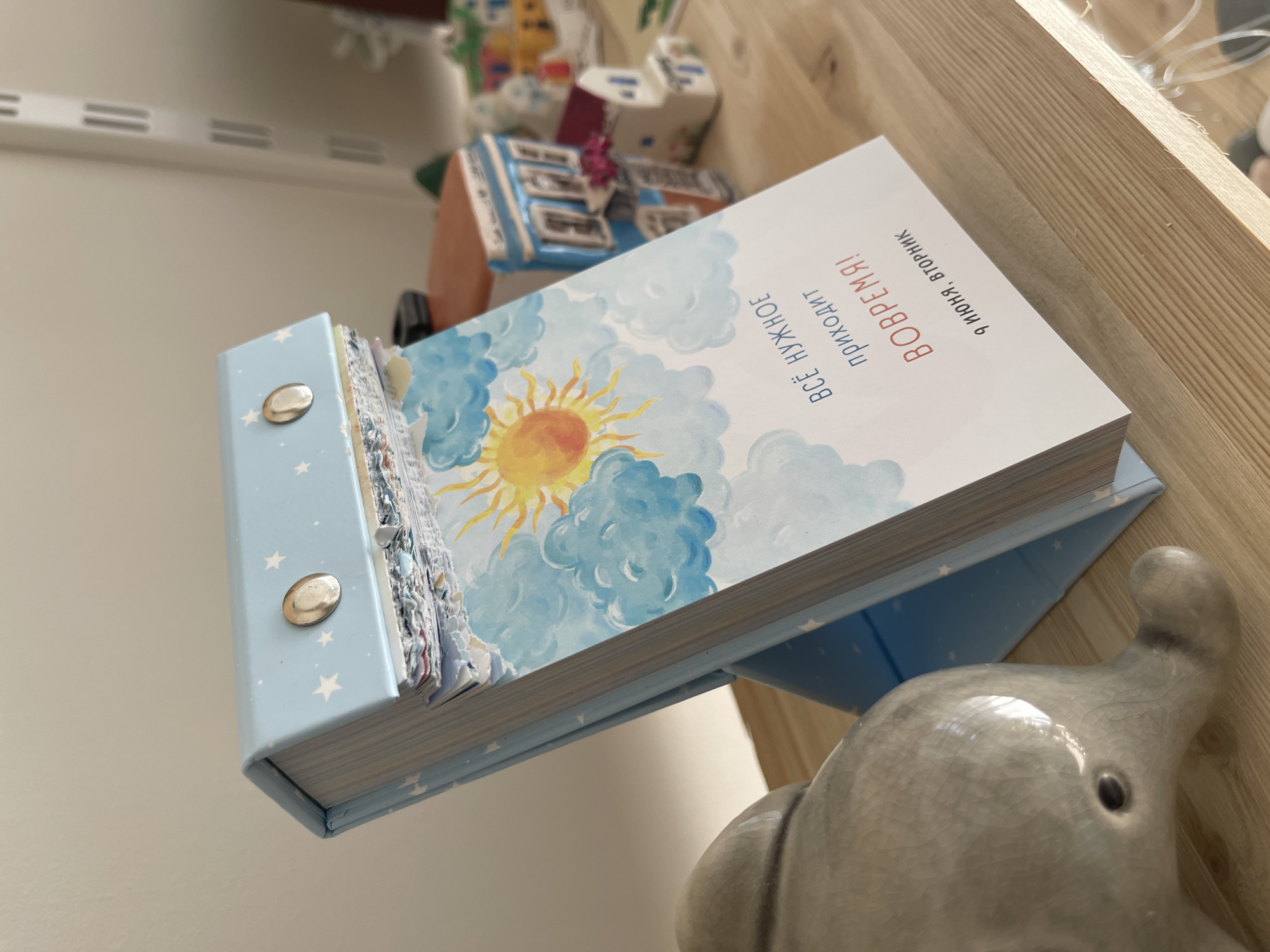 A close-up shot shows a light blue, star-patterned flip calendar standing on a wooden shelf. The calendar's front page features a watercolor illustration of a bright yellow sun shining through blue clouds. Below the illustration, in Russian text, it reads "ВСЁ НУЖНОЕ ПРИХОДИТ ВОВРЕМЯ!" (Everything necessary comes in time!) followed by "9 ИЮНЯ, ВТОРНИК" (June 9th, Tuesday). Two silver metal fasteners secure the top of the calendar. To the left of the calendar is a light gray, glazed ceramic elephant figurine. In the background, out of focus, are several miniature house decorations. The shelf itself is made of light-colored wood.