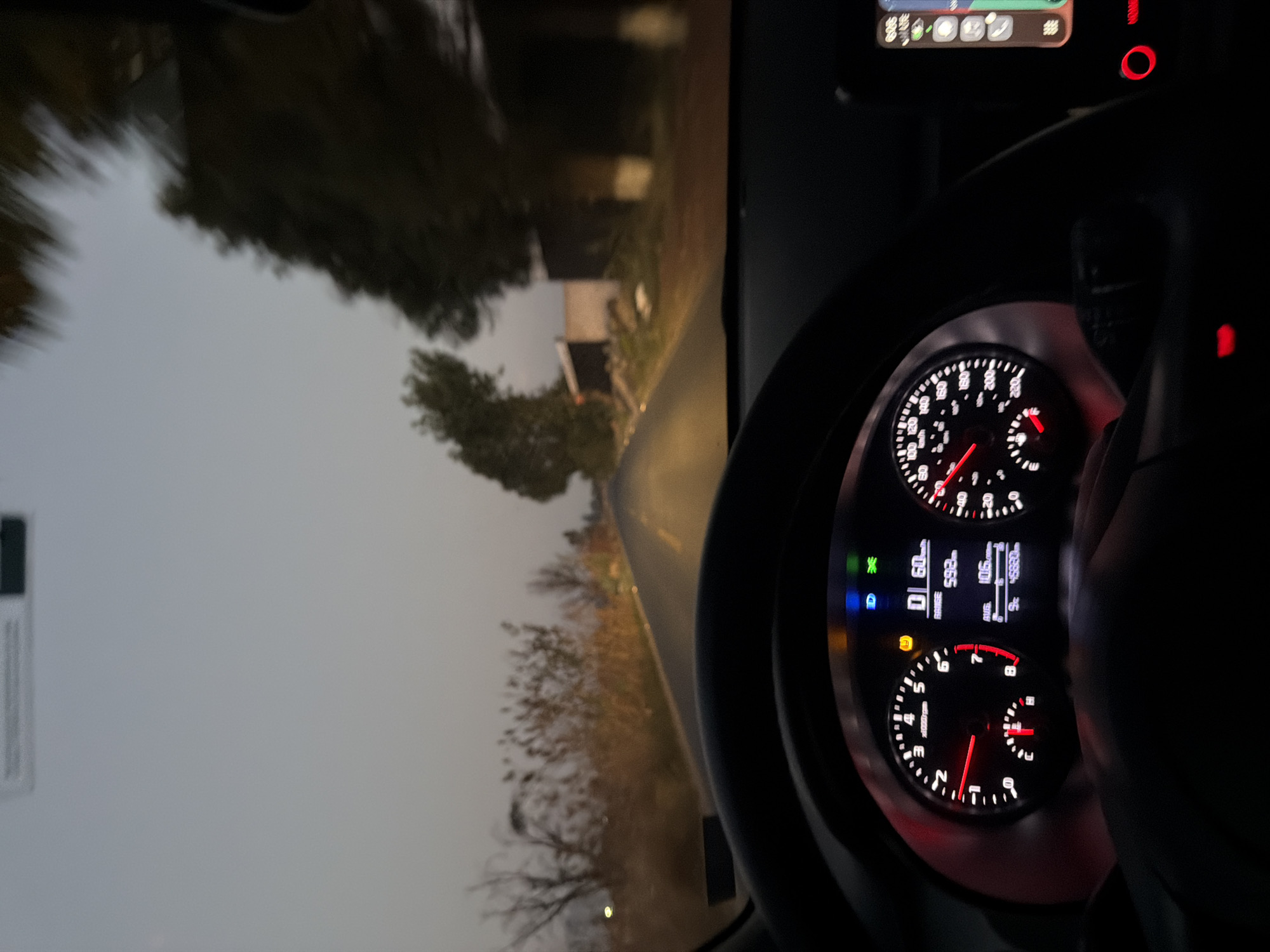 The image is taken from inside a moving vehicle, likely a car, showing the dashboard and the road ahead. The speedometer indicates a speed of approximately 60 km/h. The road is paved and appears to be winding through a somewhat rural or suburban area, with trees and a building visible on the side. The lighting suggests it is either early morning, late evening, or a cloudy day. Given the camera angle, the driver is the subject, and the action is driving. Based on the details available in the photo and with the details of the question, determining the specific location in San Pedro Cholula, Mexico, is impossible without additional information or clearer landmarks in the background.