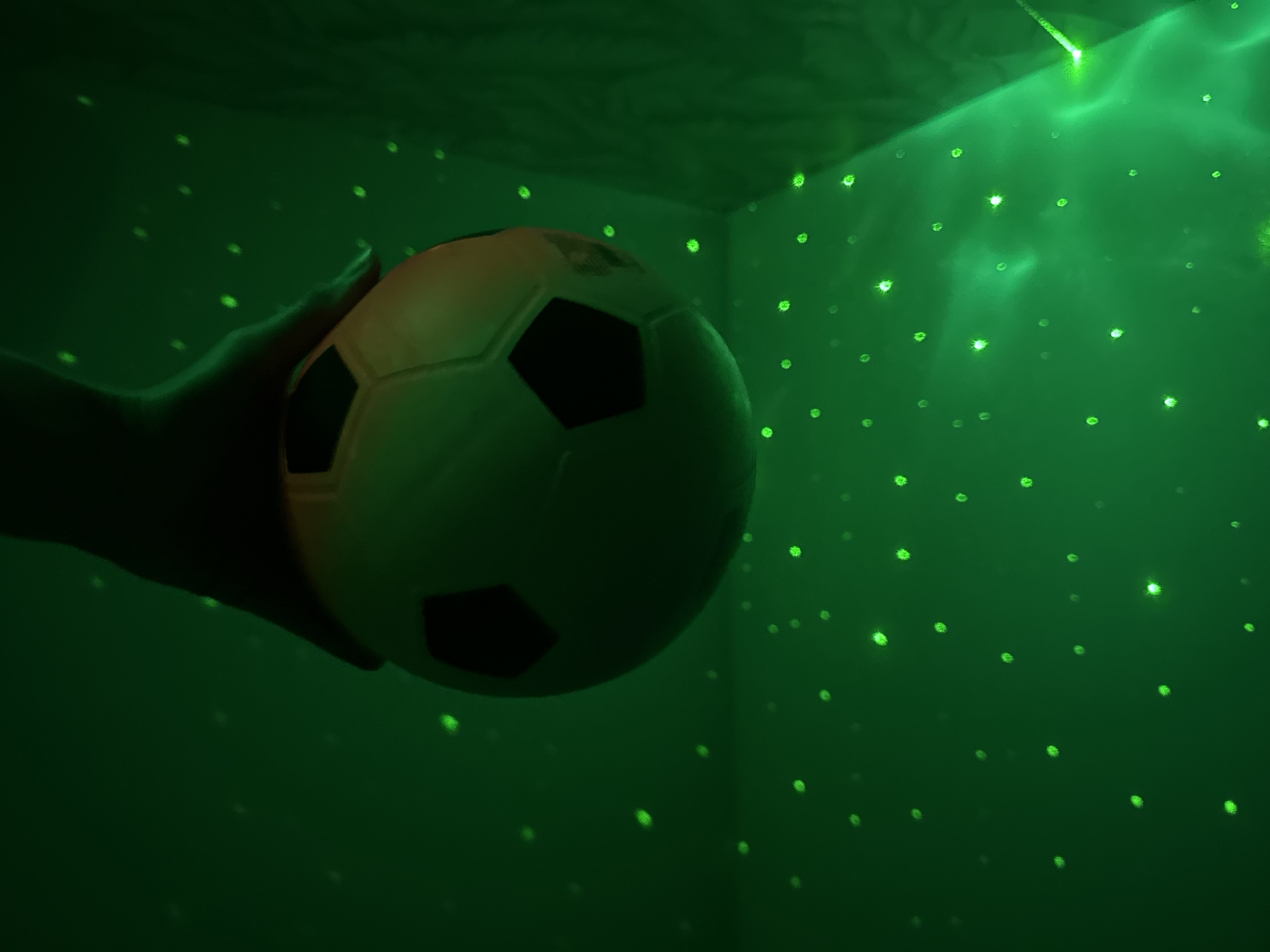 A hand holds a soccer ball in a green-lit corner. The walls and ceiling are dark green with small, bright green lights scattered across them, like stars. The soccer ball is white with black patches. The photo appears to have been taken indoors. There is no indication that the location is Прикамский, Russia.