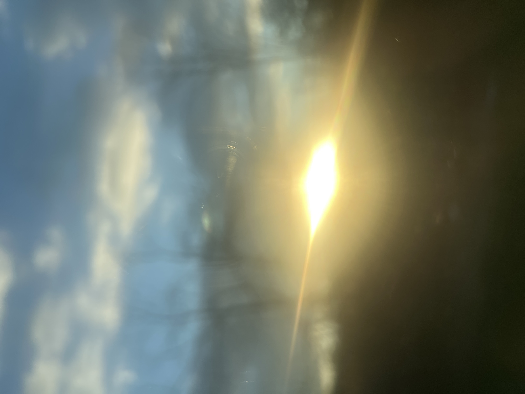 The image shows a blurred view of a sky with clouds reflected in a glass surface. A bright glare, likely from the sun, is present on the right side. The reflection gives a distorted, almost liquid appearance to the clouds. The specific location within the Town of Esopus, United States of America, cannot be determined from the visual information provided.