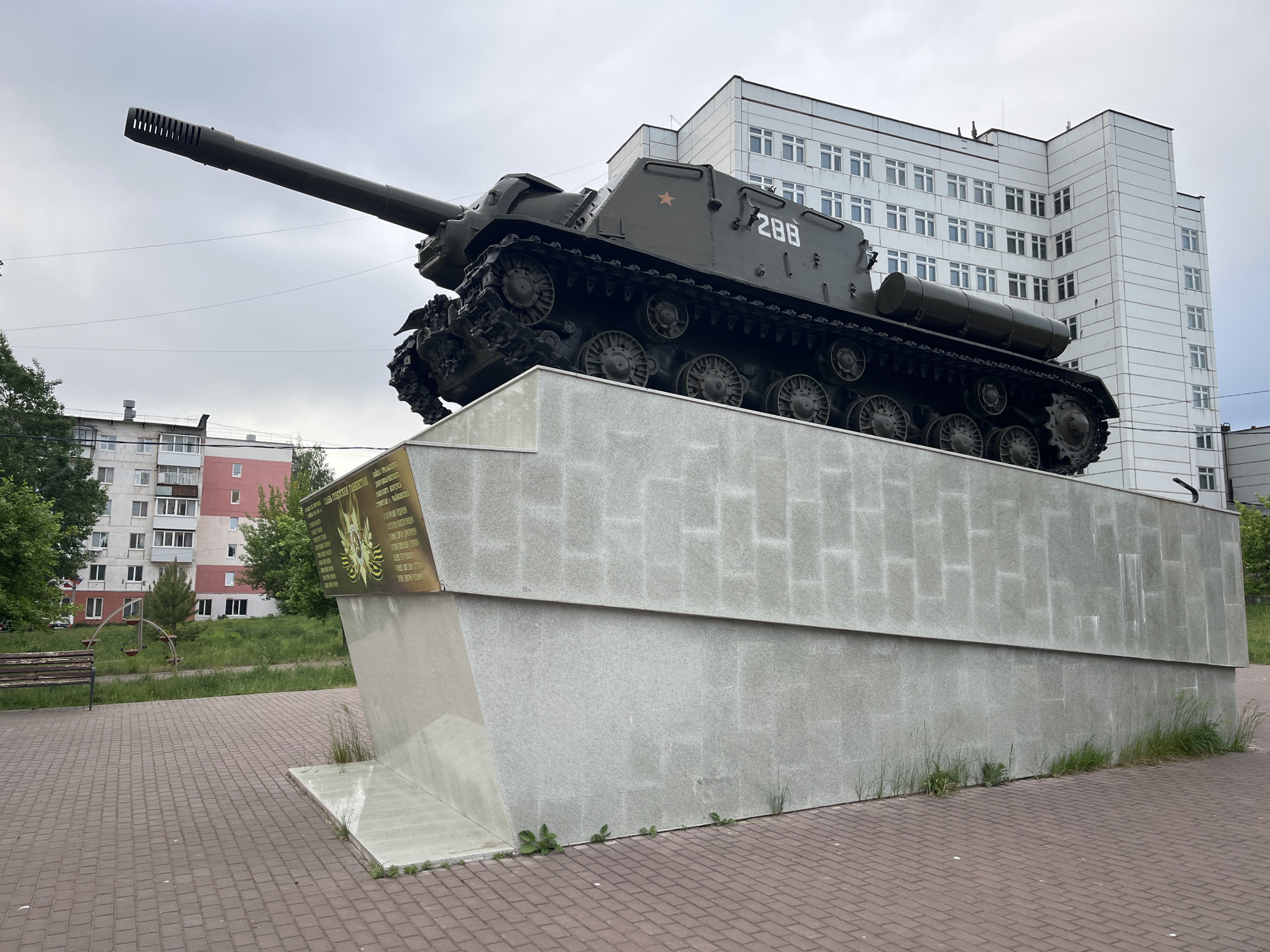 The image shows a monument featuring a Soviet-era tank mounted on a concrete pedestal. The tank is painted green and bears the number "288" and a red star. The pedestal is gray, textured in a block pattern, and slightly overgrown with grass at its base.
The background includes a multi-story white building with numerous windows and a residential building with balconies. A bench and a small playground are visible in the green space to the side. The foreground shows a paved area made of red bricks. The location is in the city of Chaikovsky, Russia.