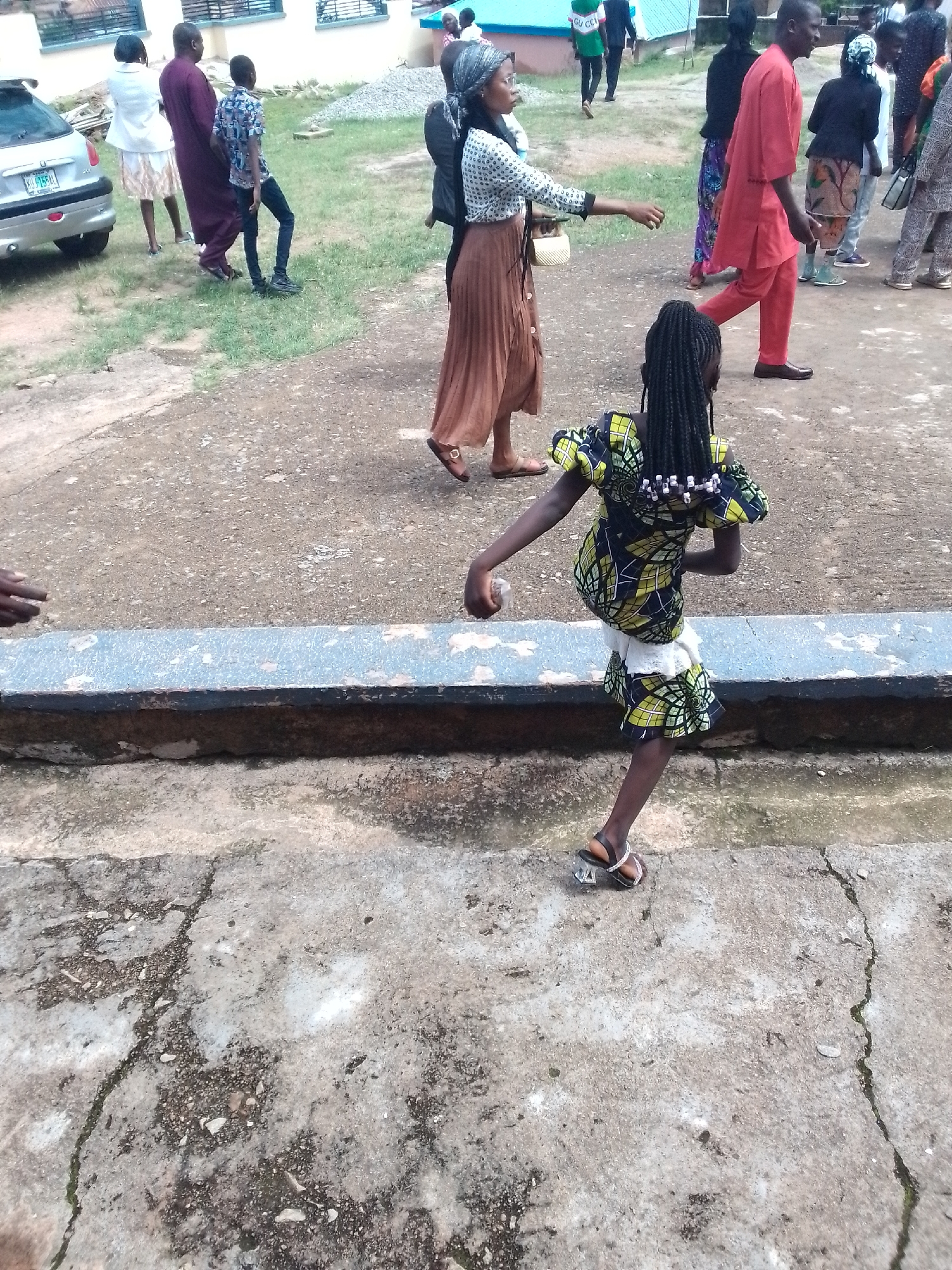 The image captures an outdoor scene in a location resembling a school courtyard or a public gathering area. The ground is a mix of cracked concrete and dirt, with a low concrete barrier running horizontally across the mid-ground. Several individuals are present, varying in age and attire.
In the foreground, a young girl with braided hair, adorned with beads, is captured mid-stride, facing away from the camera. She wears a vibrant, patterned dress and sandals. Her movement suggests she is either running or dancing.
Further back, other people are visible. To the left, a silver car is parked on a grassy area. A man in a maroon robe and a woman in a white top and skirt stand near the car, with a young boy in a patterned shirt and dark pants beside them. In the mid-ground, several individuals are walking or standing. One woman in a white dotted top and a long brown skirt is reaching out her hand. Another person in a red outfit is walking away from the camera. A woman in a black headscarf and a patterned skirt is also present. The background shows a building with windows and a light blue roof, suggesting an institutional setting. The overall atmosphere appears to be that of a community gathering or event.