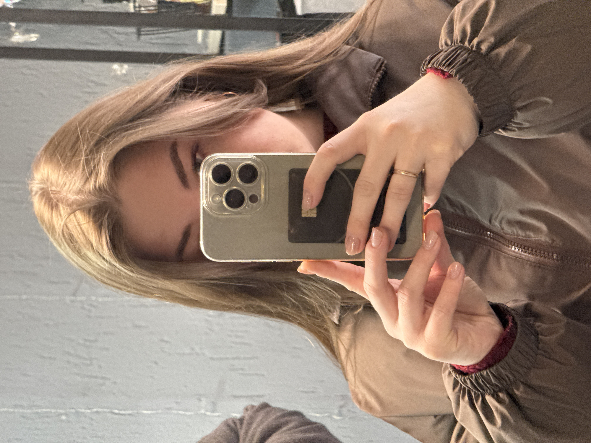 The image depicts a mirror selfie of an individual, likely female, with light brown hair, partially obscuring her face with a smartphone. Her eyes are visible, looking directly towards the phone's camera. She is wearing a dark brown or khaki-colored zippered jacket with elasticized cuffs, revealing a dark red garment underneath. Both hands hold a silver-colored smartphone, which has a clear protective case and a visible triple-lens camera setup. A dark, rectangular attachment, possibly a card holder with a silver chip, is affixed to the back of the phone case. A gold-colored ring is present on the ring finger of her right hand. Her fingernails have a light-colored polish. The background, reflected in the mirror, shows a light-colored, textured wall surface on the left and a darker, indistinct area on the right, which includes what appears to be metallic framework or shelving and some reflective decorative elements. The precise location within the city of Chaikovsky, Russia, cannot be determined from the visual evidence.