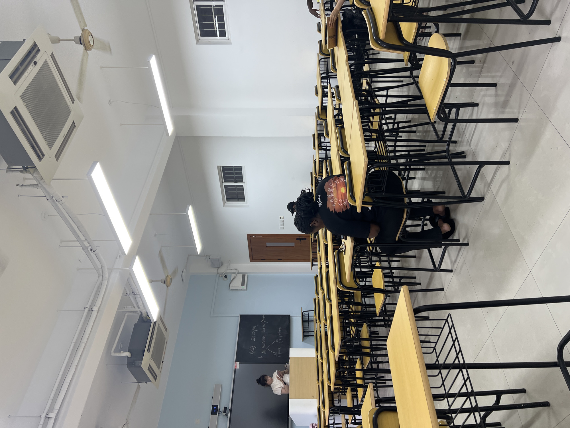 The image depicts a classroom environment, likely in a school or university setting, in Liandu District, China. The room is equipped with rows of desks and chairs, illuminated by fluorescent lighting and ceiling fans. A teacher or student stands at the front of the classroom, near a blackboard displaying some writing. Several students are seated at desks, some appearing to be engaged in learning activities. The room has a clean and organized appearance, with white walls and a tiled floor. The overall atmosphere suggests a typical educational setting.