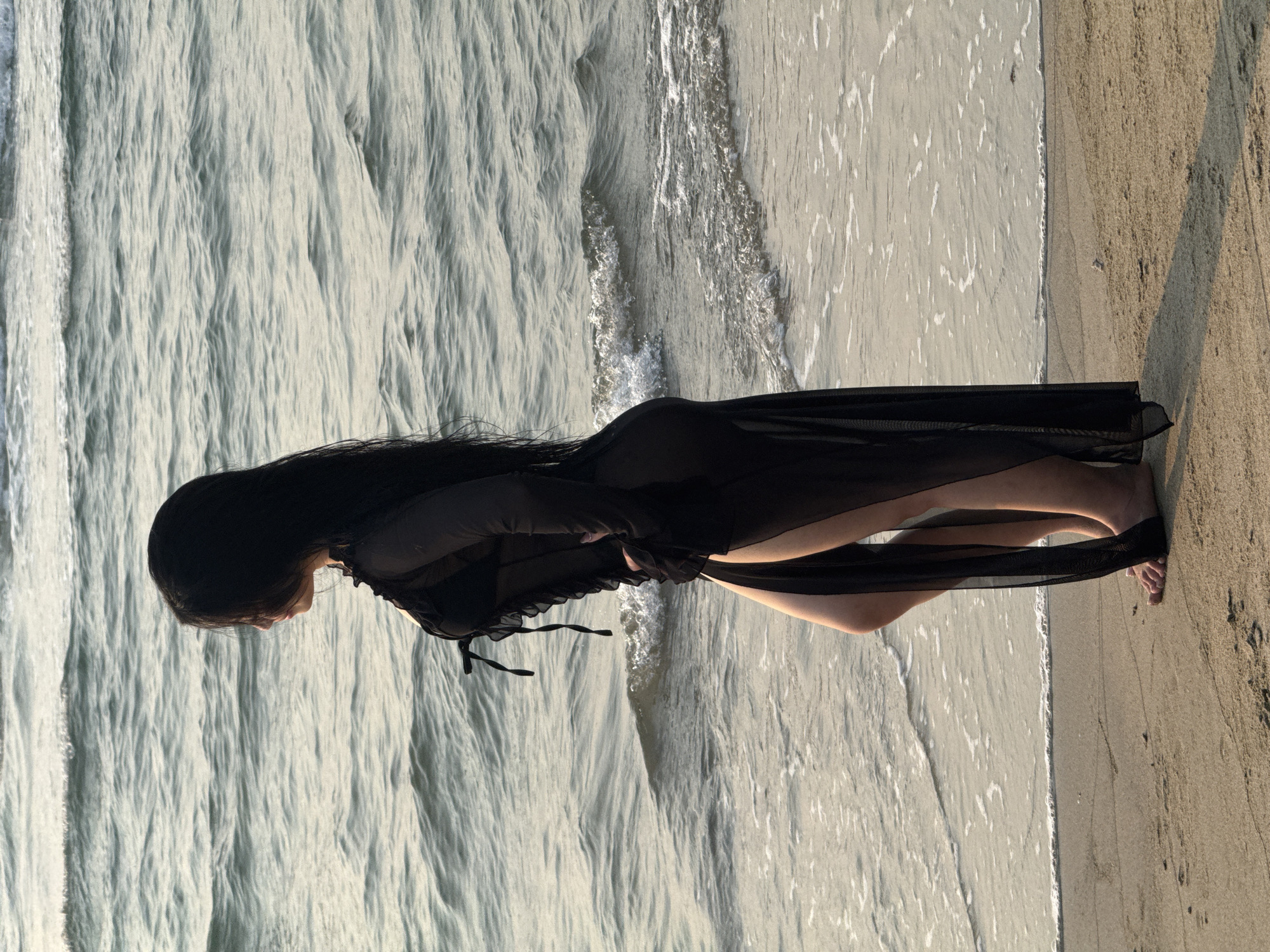 A woman in a black sheer dress stands on a sandy beach at the edge of the water. The water is a light grey-green color with small waves. The sand is light brown. The woman has long, dark hair.
The location appears to be a beach in Đà Nẵng, Vietnam.