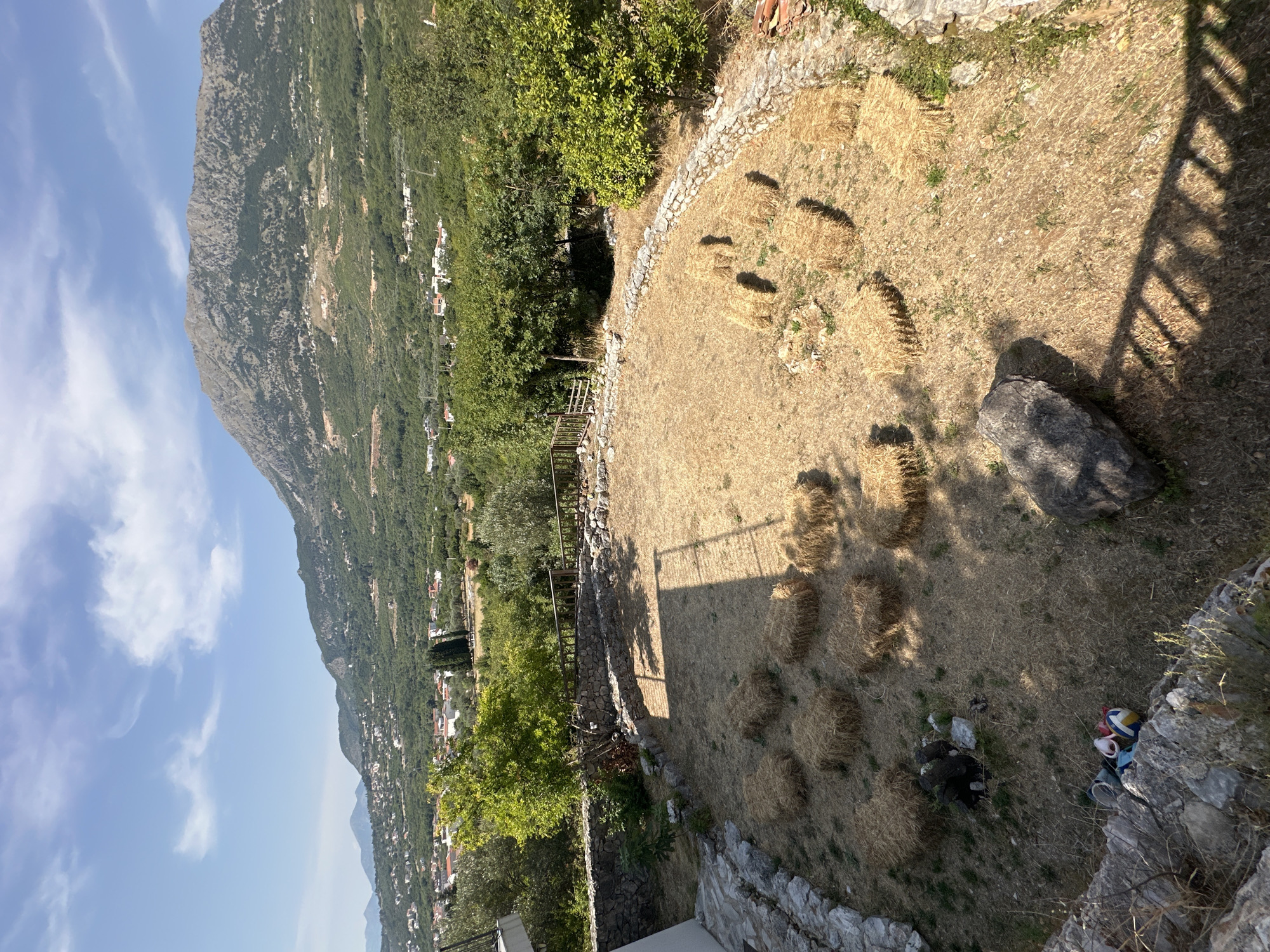 In Pečurice, Montenegro, a sun-drenched, open-air gathering space features several bales of hay arranged in a circular fashion on dry, sparse grass. The space is bordered by a low, rustic stone wall, and a wooden fence. Beyond the immediate area, the landscape transitions into lush green trees and vegetation, with a village nestled into the hillside. In the background, a large, rugged mountain dominates the horizon under a clear blue sky with scattered white clouds. A white building is visible on the left edge of the frame, and some scattered debris, including a yellow and blue ball and a red object, are visible in the lower left corner.