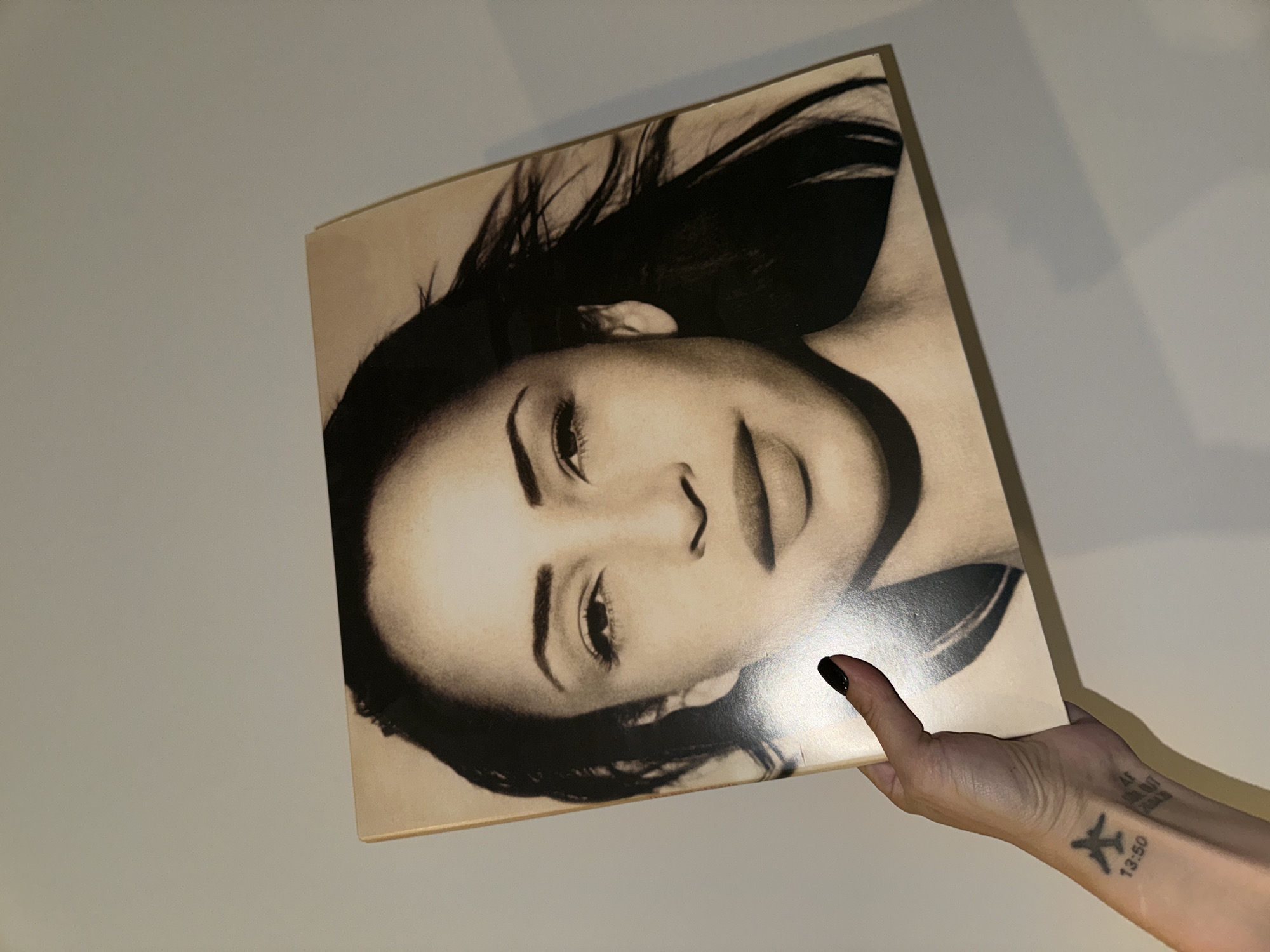 A close-up, eye-level shot captures a person's hand holding up a large, square-shaped object, likely a vinyl record album cover or a print, against a plain, light-colored wall in what appears to be an indoor setting in Moscow, Russia. The object is oriented diagonally, tilted upwards from the bottom right.
The most prominent feature of the object is a sepia-toned or black and white portrait of a woman's face, rotated approximately 90 degrees clockwise. Her dark hair flows around her head, and her gaze is directed upwards (or to the viewer's right). She has high cheekbones, defined eyebrows, and a slightly open mouth. The artwork style emphasizes shading and contrast, giving the portrait a timeless, artistic quality.
The hand holding the object is positioned at the bottom right corner. The person has black nail polish on their index finger and thumb. Visible on the inner wrist and forearm are several black tattoos. A clear tattoo depicts a small airplane silhouette, with the numbers "13:50" directly beneath it. Above the airplane, partially obscured by the album cover and the angle of the arm, are other tattooed characters that appear to be "AF", "47", and possibly "2018". The lighting is even, casting subtle shadows on the wall behind the hand and the object. No other subjects or activities are visible.