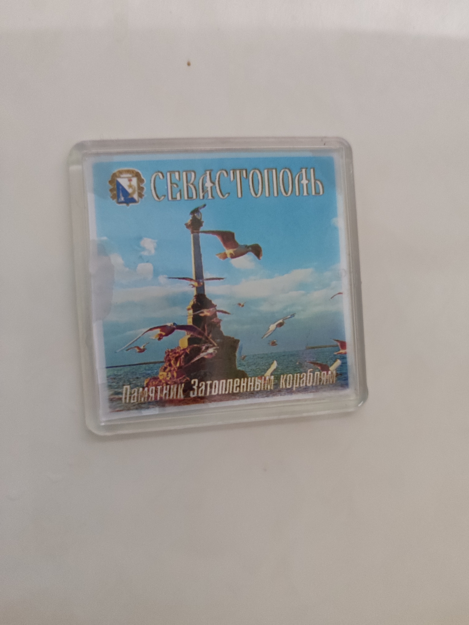 The image shows a magnet with a picture of the Monument to the Scuttled Ships in Sevastopol. The monument is depicted in the foreground, with several seagulls flying around it. The sea and the sky are visible in the background. Text on the magnet reads "СЕВАСТОПОЛЬ" and "Памятник Затопленным кораблям". This image is a souvenir and is not related to Bondarivka, Ukraine.