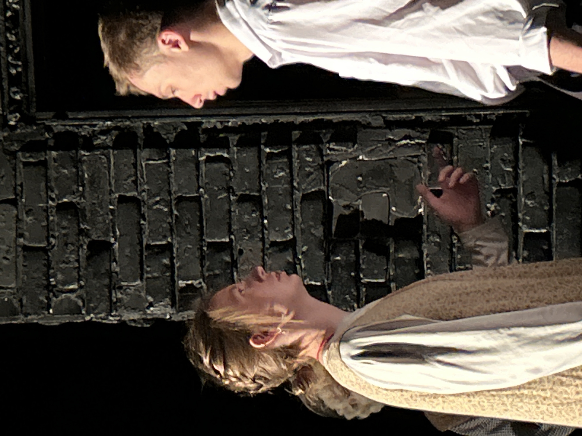 Two individuals are visible within the frame, positioned against a dark, brick-like structure. The person at the top is fair-skinned with short, light-colored hair, wearing a white shirt. Below, the other individual, also fair-skinned, has long, light brown hair styled with braids. They are clad in a light-colored, patterned vest over what appears to be a white garment. The upper individual's hand is raised near the brick structure. The background is a dark void, suggesting an indoor setting, possibly a theatrical stage.