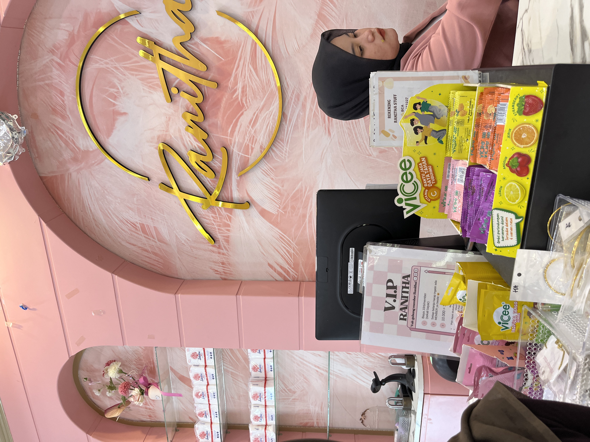 This is a medium shot of a young woman wearing a black hijab and a pink jacket, seated at a counter in what appears to be a beauty or cosmetic store. The store has a predominantly pink color scheme, with arched decor and feathered wallpaper in the background. A large, ornate golden cursive sign that reads "Ranitha" is prominently displayed on the wall behind the woman.
In front of the woman on the counter, there is a computer monitor and a display stand filled with small packets of "V₁cee" brand vitamin C tablets in various fruit flavors. To the left of the display stand, there's a small basket containing what seem to be bracelets and other jewelry items. A sign on the counter, partially obscured by a display, reads "V.I.P RANITHA" and lists membership benefits. The overall ambiance suggests a retail environment focused on beauty products and accessories, likely catering to a younger demographic. The location is identified as Lhokseumawe, Indonesia, based on context from the user.