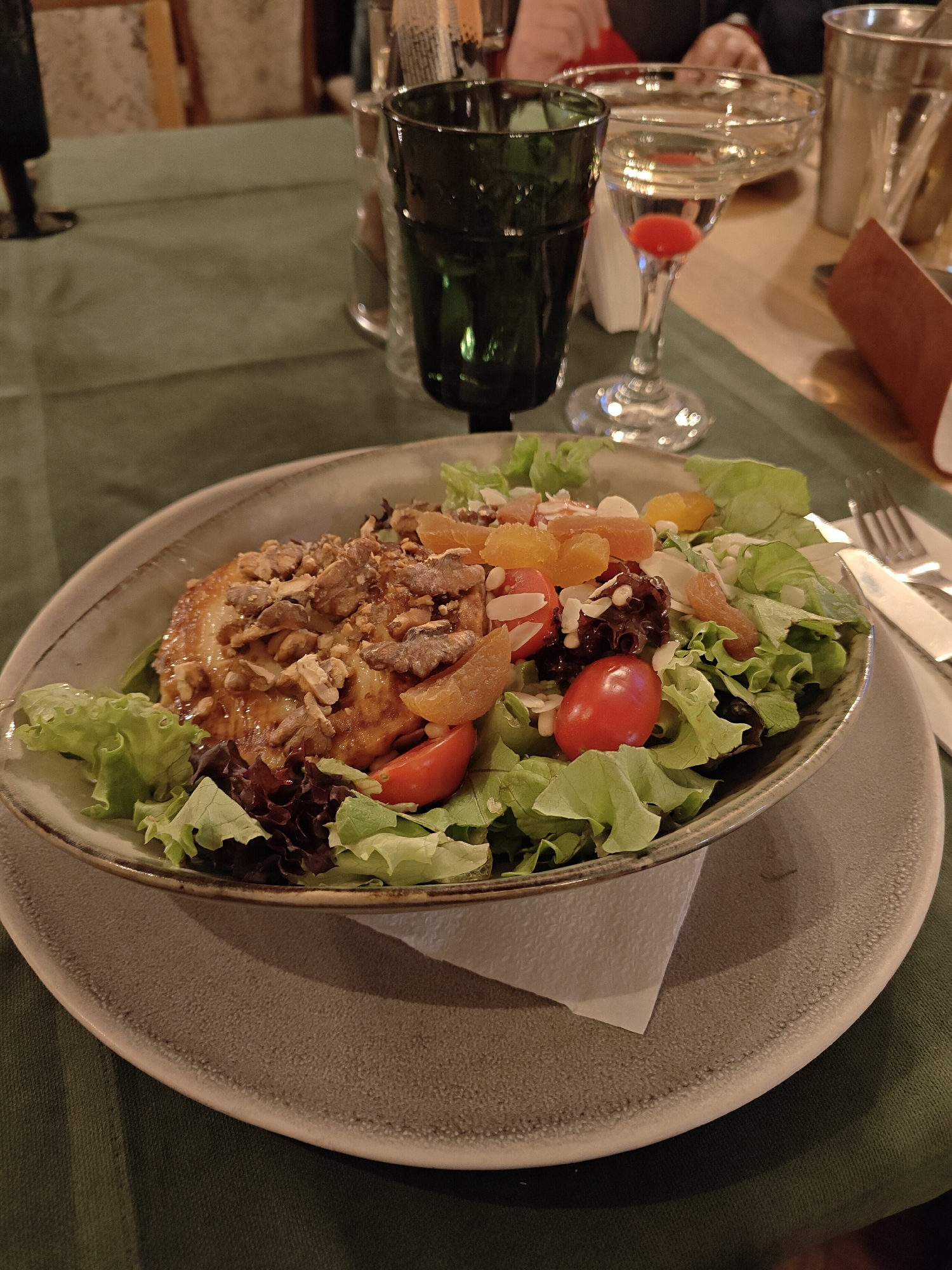 A salad is centered on a table in a room with multiple tables and people, though Baldevo, Bulgaria, cannot be confirmed based on this image. The salad contains lettuce, red tomatoes, orange fruit pieces, almonds, and walnuts atop a large piece of bread. The salad sits in a grey bowl which is centered on a grey plate and a white napkin is placed underneath the bowl. To the right of the bowl are a fork and knife. Behind the salad are two drinking glasses, one green and filled and another containing liquid with a red cherry. The table is covered with a green tablecloth and there are chairs and people visible in the background.