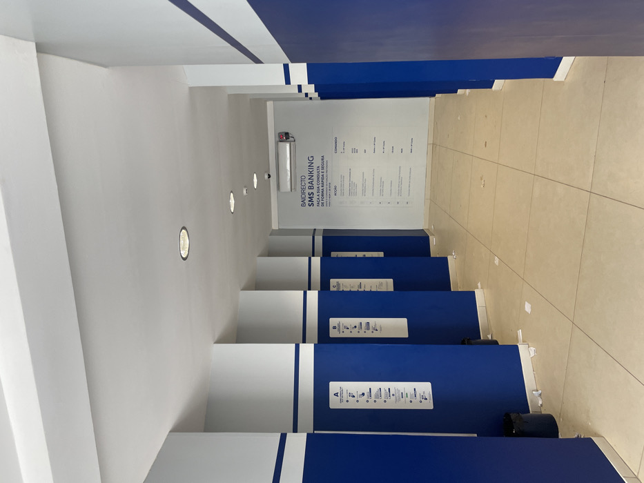 The image displays an empty, well-lit indoor corridor or service area, characteristic of a banking facility, located in Luanda, Angola. The space is dominated by a clean and modern blue and white color scheme.
The walls and ceiling are white, accented with bold horizontal and vertical royal blue stripes. The floor is tiled with light beige or cream-colored rectangular tiles, showing minimal wear.
Along the right side of the corridor, a series of individual service stations or cubicles are arranged, providing a sense of privacy. These partitions are primarily blue with white tops. Each cubicle features a white informational sign, visibly labeled "A", "B", and "C" (from the foreground to the background). These signs contain pictograms and brief text, likely instructing users on how to access or utilize banking services. A small black waste bin is present near the first cubicle on the right.
At the far end of the corridor, a prominent white wall serves as an information display. It features a security camera mounted near the top and a large poster or screen with text. This text advertises "BAIDIRECTO SMS BANKING" and states in Portuguese: "FAÇA A SUA CONSULTA DE FORMA RÁPIDA E SEGURA" (meaning "MAKE YOUR CONSULTATION QUICK AND SAFE"). Below this, columns of information are visible under the headings "ACÇÃO" (ACTION) and "COMANDO" (COMMAND), detailing specific SMS banking commands and their corresponding actions.
The area is artificially illuminated by several circular recessed lights embedded in the ceiling. No people are present, giving the impression of either off-hours or a quiet period of operation. The environment is orderly and functional, designed for efficient customer interaction within a financial institution.