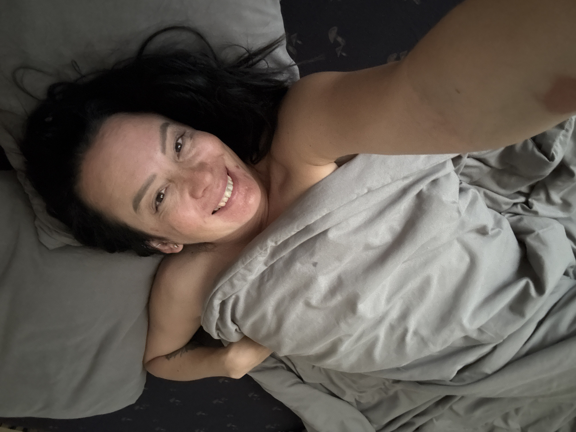 A woman is lying in bed, taking a selfie. She has dark hair and is smiling. She is partially covered by a grey blanket. The bed has grey sheets and a dark patterned bedspread. The location is potentially inside a residence, but there is no verifiable information from the image to confirm that it is in Chaikovsky, Russia.