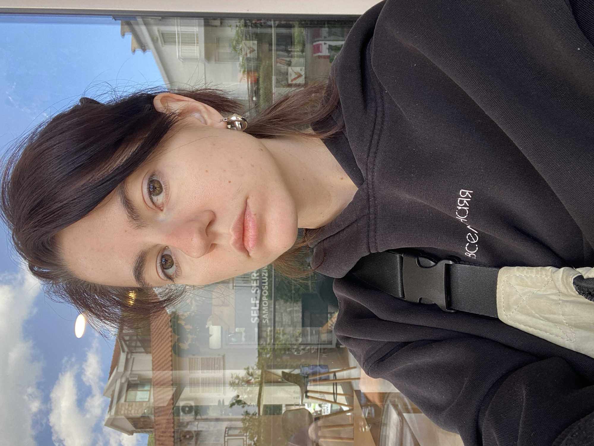 A medium close-up shot, rotated counter-clockwise, features a young woman with light skin, brown hair with lighter streaks, and brown eyes, looking directly at the camera with a slightly pensive expression. She is wearing a black hoodie, with some white text visible on the upper left side. A light-colored bag with a black buckle strap is slung across her body.
The background is a window reflecting an exterior scene. The reflection reveals a multi-story building with a red-tiled roof, windows, and what appears to be air conditioning units. A blue sky with some clouds is also reflected in the window. Reflected in the window is a cafe interior, with tables and chairs. The reflection indicates an urban location. The text "SELF-SER" and "SAMOPOSLUZI" are visible in the reflection as well. The location is Unknown, Unknown.