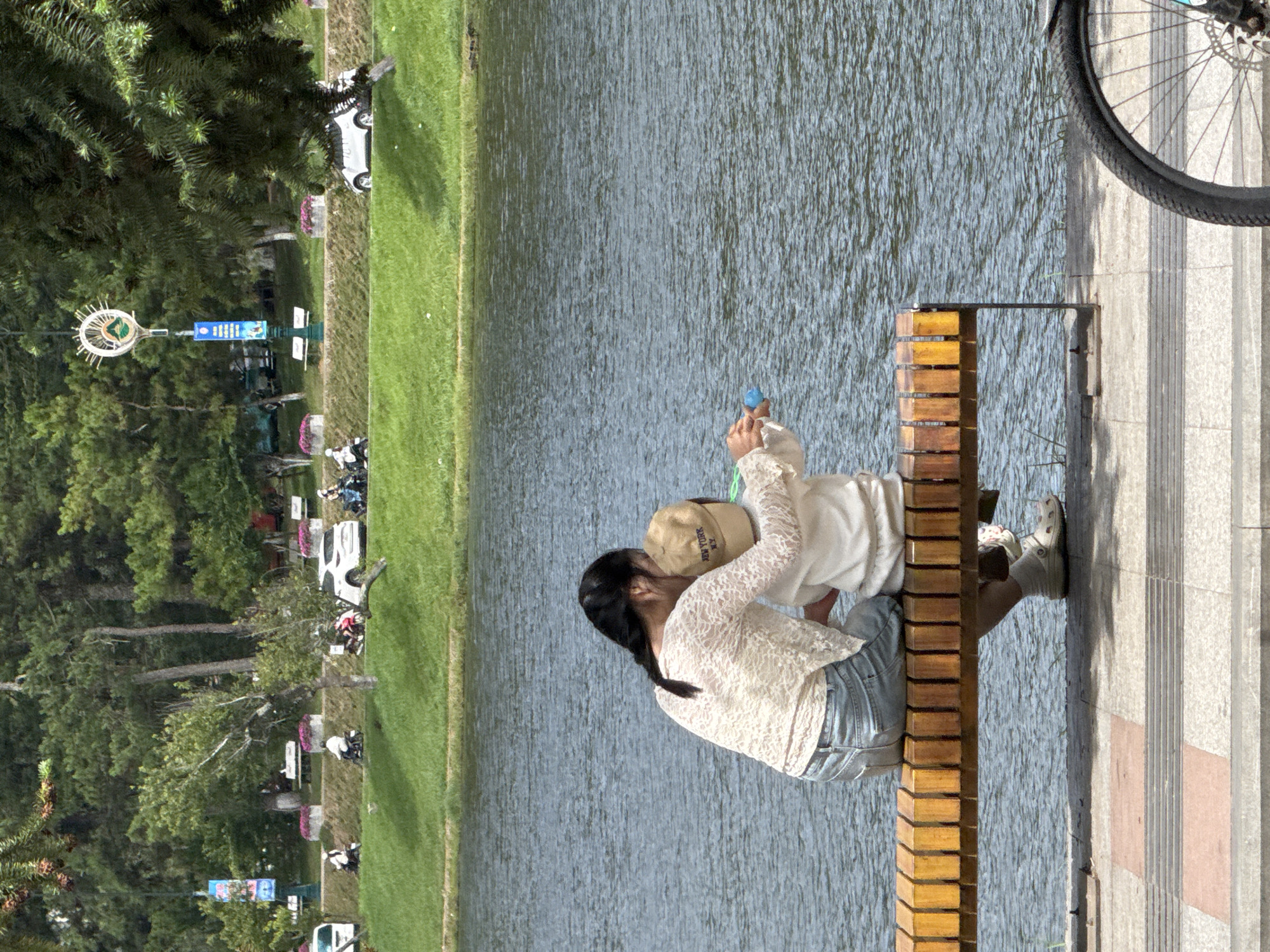 The image depicts a serene waterside scene in Da Lat, Vietnam. In the foreground, a woman and a child are seated on a wooden bench, facing a body of water. The woman, wearing a white lace top and denim jeans, has her arm around the child, who is dressed in a white outfit and a tan baseball cap. The child appears to be holding a blue object. To the right of the bench, the front wheel of a bicycle is visible.
The background features a lush green hillside dotted with trees. Further back, a road runs parallel to the hillside, with several white cars and motorcycles parked or in motion. Benches and decorative planters with pink flowers line the edge of the road. A tall lamp post with a circular decorative element and a blue banner stands near the trees. The water in the foreground is calm with visible ripples. The overall atmosphere is peaceful and appears to be a public park or promenade area.