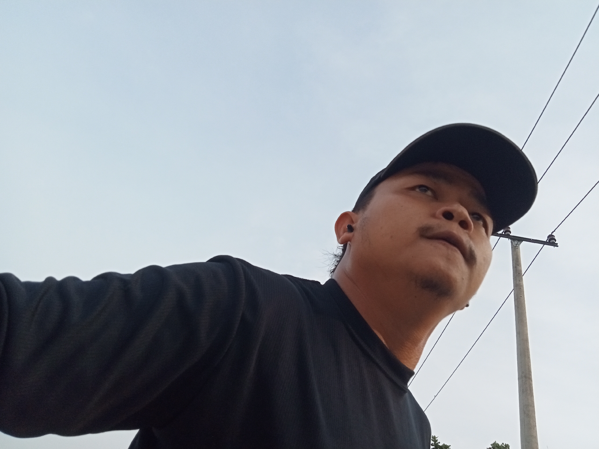 A low-angle, wide shot captures a person wearing a black t-shirt and a black baseball cap. The person is looking upwards and to the right. Their face is partially obscured by the cap and the angle of the shot. A utility pole with wires is visible in the background, against a pale sky. The image is taken outdoors, likely in the city of Teluk Kenidai, Indonesia, as indicated by the presence of the utility pole and wires common in the area. The lighting suggests it is daytime, possibly overcast.