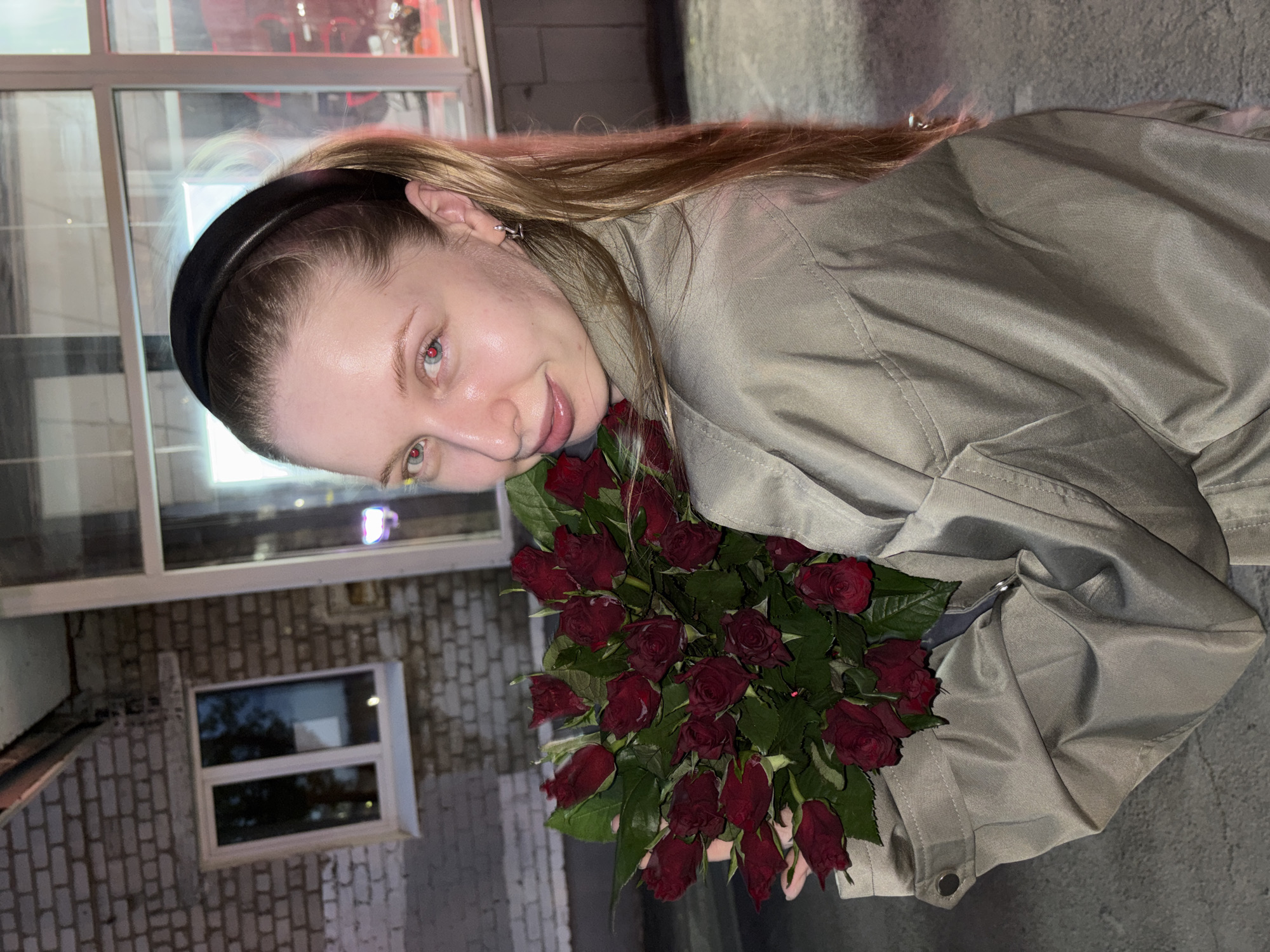 A woman is holding a bouquet of dark red roses, possibly outdoors. She has fair skin, blond hair pulled back with a black headband, and is wearing a beige, buttoned shirt or jacket. The background includes what appears to be a brick building with a window, and a glass-fronted structure possibly reflecting red lighting. The location is potentially Perm, Russia.