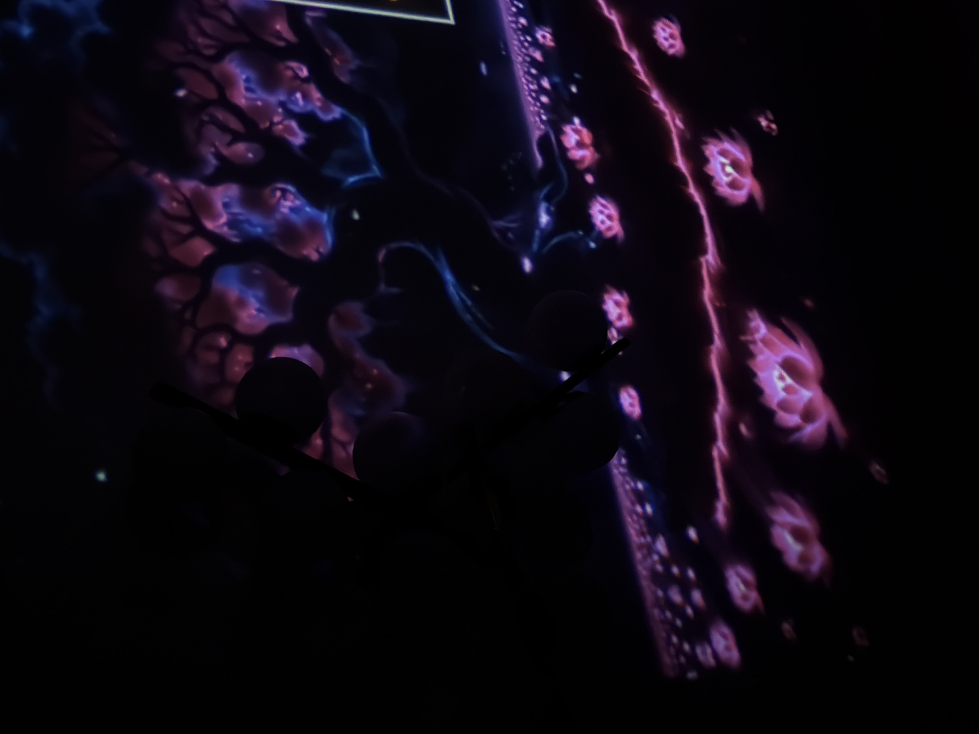 The image shows a dark scene with a projection of a fantasy landscape featuring stylized trees with pink and blue foliage against a dark background. In the foreground, there are rows of glowing pink lotus-like flowers. A dark silhouette of a hand holding a stylus or pen is partially obscuring the left side of the image. The setting appears to be indoors, possibly an exhibition or performance space. There is no indication of the city of Tbilisi, Georgia.