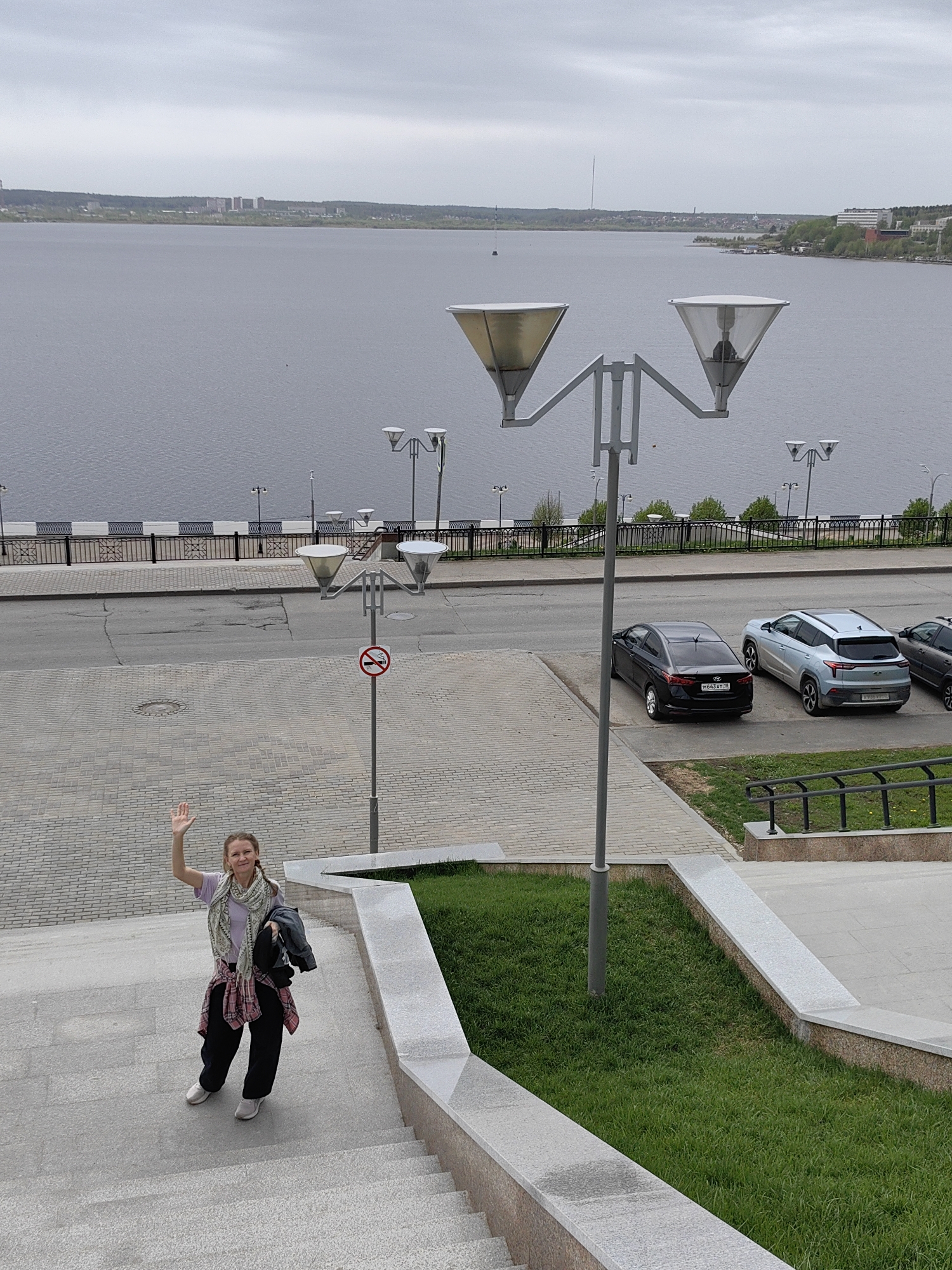 A woman is standing on a staircase, waving her right hand towards the viewer. She is dressed in a light purple t-shirt, black pants, and white sneakers, and she has a patterned scarf and a plaid shirt around her neck and waist, respectively. She is also holding a black jacket.
Behind the woman, the staircase leads down to a paved area with street lights, cars parked along the side, and a view of a large body of water with a cityscape in the distance. The water appears calm under a cloudy sky.
There is a "no smoking" sign attached to one of the street light poles. Two cars are parked in a parking area: a black Hyundai and a light blue Moskvich. The location is Izhevsk, Russia.