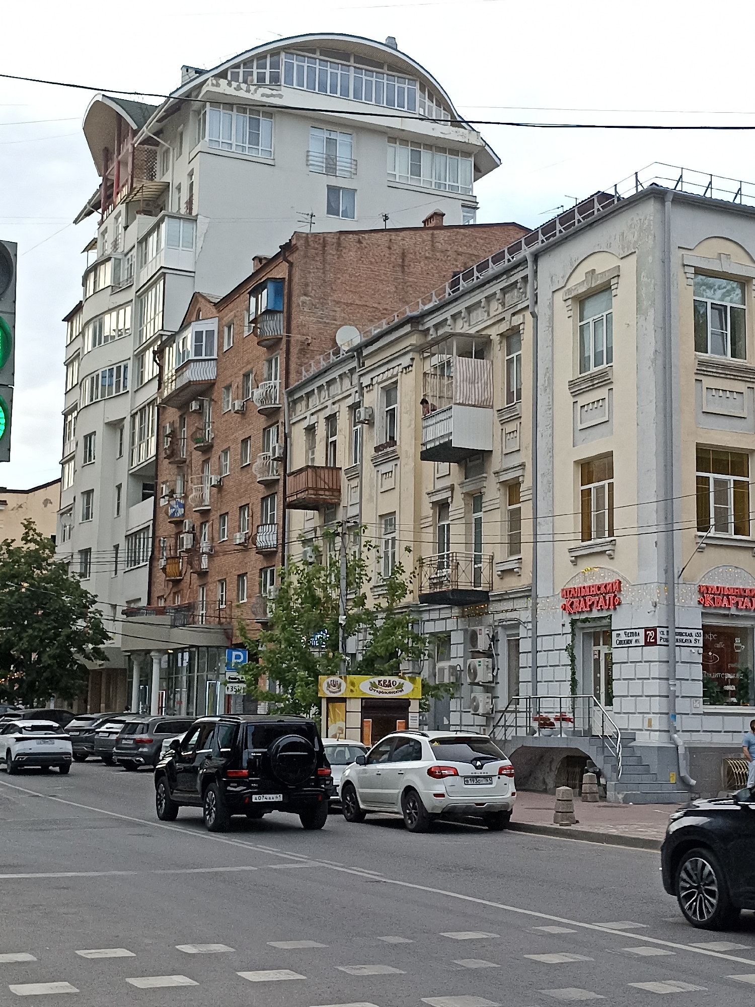 The image depicts a street scene in Rostov-on-Don, Russia, likely in the area of Chekhov Street and Pushkinskaya Street. The foreground features a paved road with white crosswalk markings and several cars driving or parked. A black SUV with a spare tire on the back is prominent in the center, displaying the license plate A074AA 161. To its left, a white SUV is parked, and further down the street, a row of parked cars is visible.
The background is dominated by multi-story buildings lining the street. On the left, a modern white building with a curved roof and numerous windows stands out. Adjacent to it is a taller building with a brick facade and balconies. To the right, a lighter-colored building with decorative architectural elements and arched windows is visible. Signage in Russian is present on some of the buildings, indicating commercial establishments like cafes and shops. For example, signs for "ИТАЛЬЯНСКИЙ КВАРТАЛ" (Italian Quarter) are clearly legible. The sky is overcast. The overall impression is of an urban environment with a mix of architectural styles.