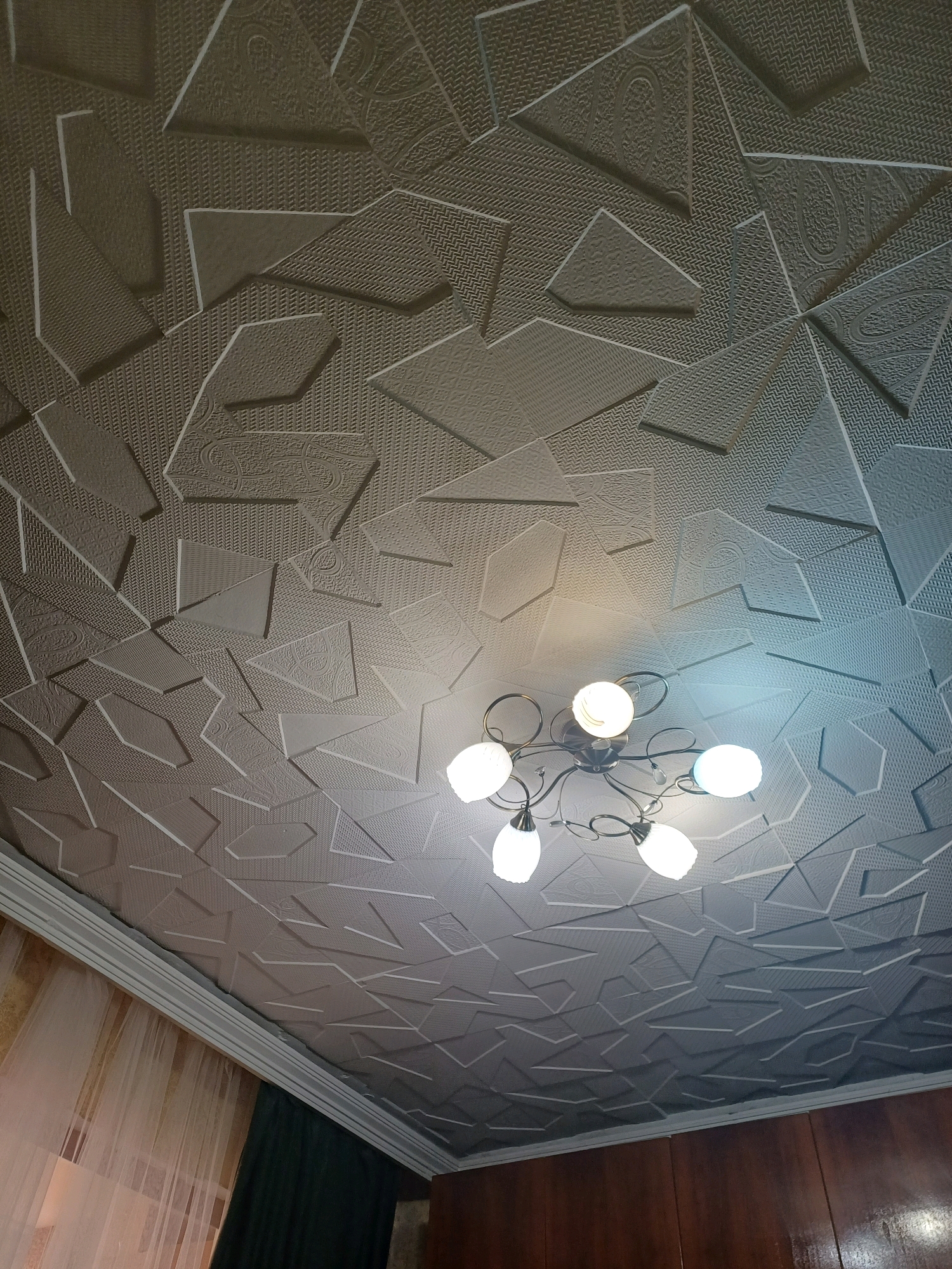 The image shows a textured ceiling with a geometric pattern of embossed shapes in various orientations. The ceiling is lit by a chandelier with five white globes and a metallic base with swirling arms. To the left of the ceiling, a portion of a sheer curtain and a dark green curtain is visible, suggesting a room interior. The style of the ceiling suggests it is likely in a residential setting, possibly a bedroom or living area. The lighting is warm, with some of the globes casting a yellowish glow and others appearing brighter, possibly due to lens flare or the angle of the camera. The overall impression is that of an interior shot of a home, with a focus on the ceiling's unique design and the overhead lighting fixture. There are no people visible in the image.