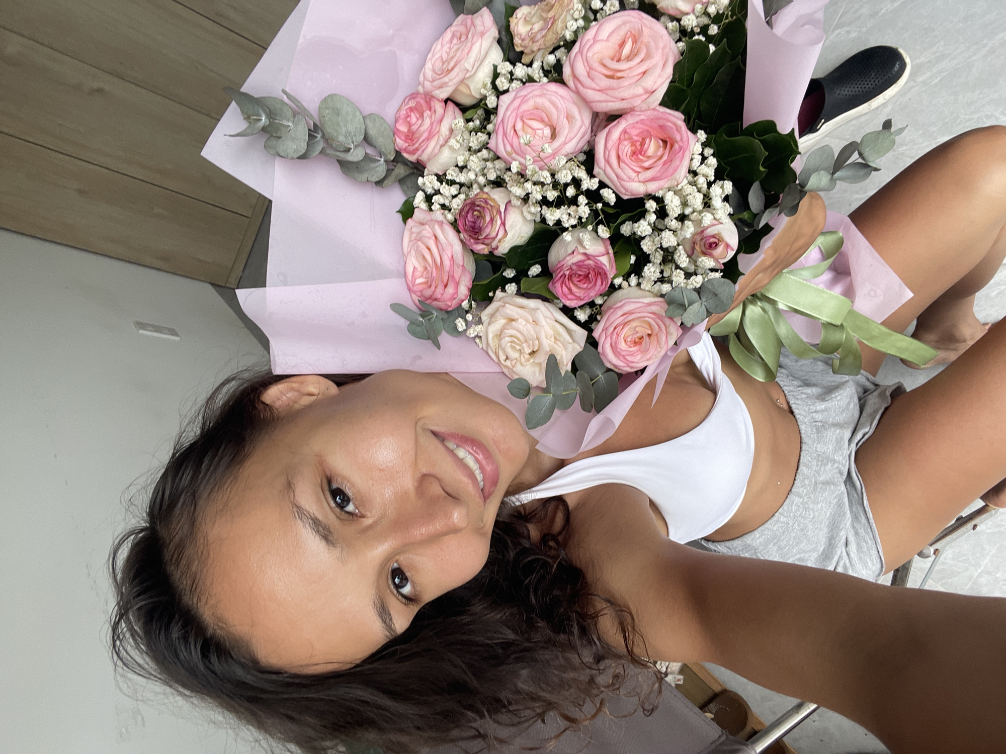 A young woman with dark, curly hair is holding a bouquet of pink and white roses wrapped in pale pink paper. She is wearing a white sports bra and grey shorts. She is smiling at the camera. The bouquet is adorned with eucalyptus leaves and baby's breath. A black clog is visible near the bottom right of the image. The background shows a light-colored wall with a wooden accent. The scene appears to be indoors, possibly in a hotel room or residential setting in Mũi Né, Vietnam.