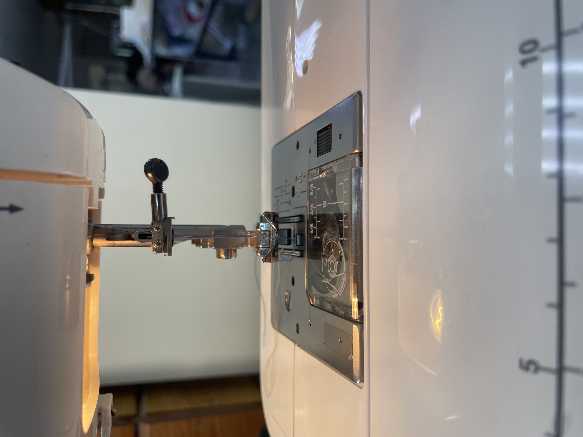 A close-up, eye-level, indoor shot shows the needle assembly of a white sewing machine. The needle, threaded with light blue thread, is positioned directly above the metal throat plate. A small black knob, likely part of the presser foot lifter, is visible to the right of the needle. The throat plate has markings for measurements, including "1/8", "3/8", and "5/8". A clear plastic cover over a bobbin winder is also visible on the throat plate. The machine is illuminated by a warm light source, casting reflections on the glossy white surface of the sewing machine. In the blurred background, what appears to be a shelf unit with some items can be seen. The scene is devoid of any people. The location is not specified in the image or the provided context.