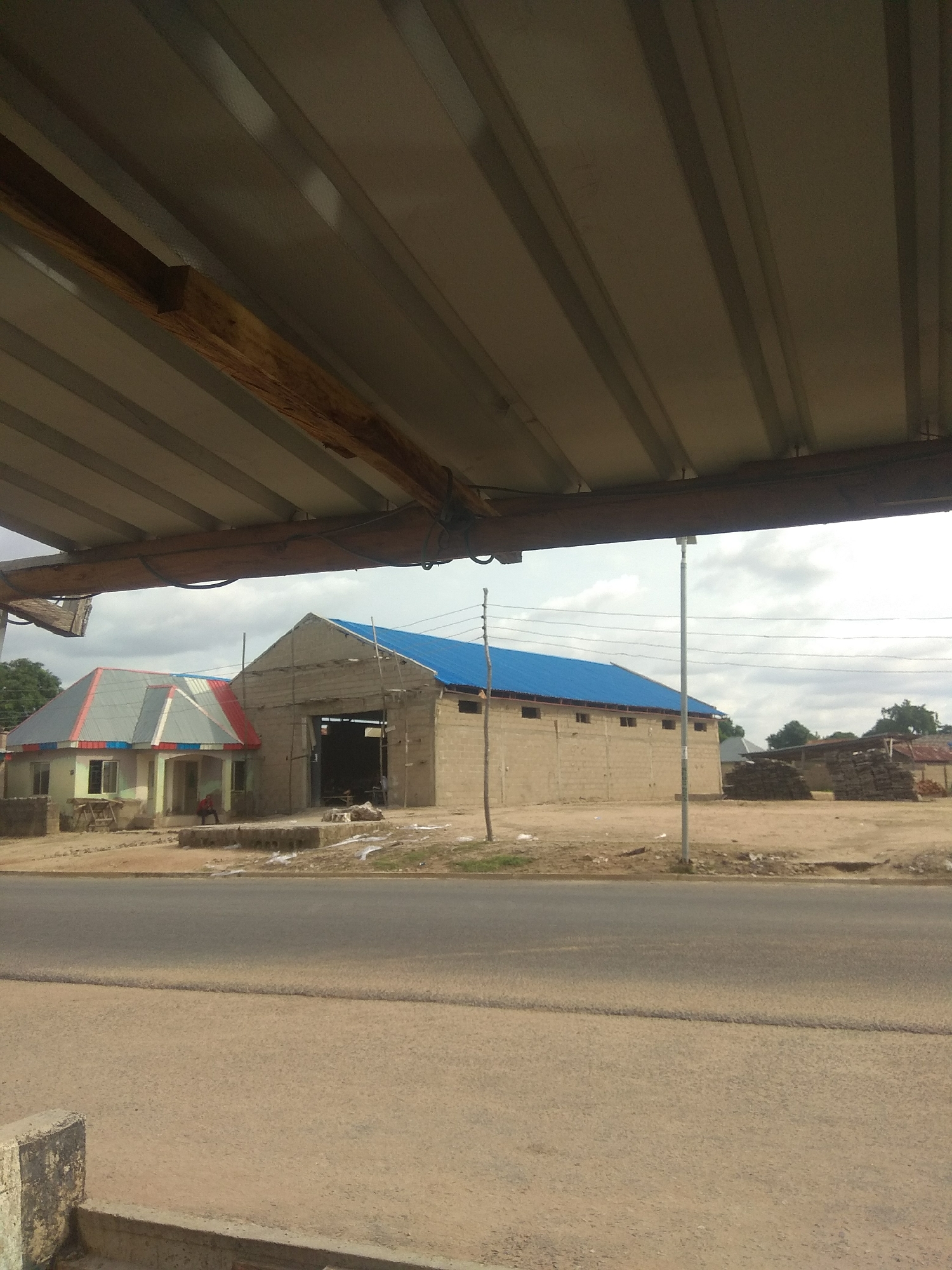 The image shows a street scene in Jalingo, Nigeria, with a focus on buildings under construction and in use. In the foreground, a gray asphalt road is visible, with a concrete curb on the left side. Above the road, a row of buildings is situated. To the left, a smaller building with a multi-colored corrugated metal roof, featuring red, white, and blue sections, is partially visible. This building has windows and appears to be inhabited, as a person is seated outside. Adjacent to it, and slightly to the right, is a larger, more industrial-looking structure. This building is constructed of cinder blocks and has a bright blue corrugated metal roof. The front of this building features a large open entrance, suggesting it is a warehouse or workshop, with scaffolding visible near the roofline. The ground around the buildings is a dusty, sandy terrain. In the background, more structures, including what appear to be stacks of lumber or building materials, are scattered, along with trees and a cloudy sky. A light pole stands near the blue-roofed building. The image is taken from beneath an overhanging corrugated metal roof structure, with a wooden beam visible, suggesting the viewpoint is from under a shelter or awning.