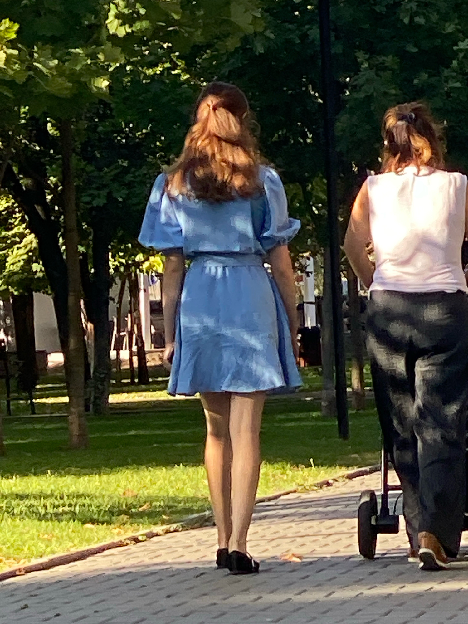 A full body rear shot captures two women walking on a paved pathway in a park. The woman on the left wears a light blue dress and walks ahead of the woman on the right. The woman on the right wears a light pink top and dark pants, pushing a stroller. The park features lush green grass, trees, and sunlight filtering through the foliage. The pathway is made of grey interlocking pavers. The scene appears to be in Chișinău, Moldova.