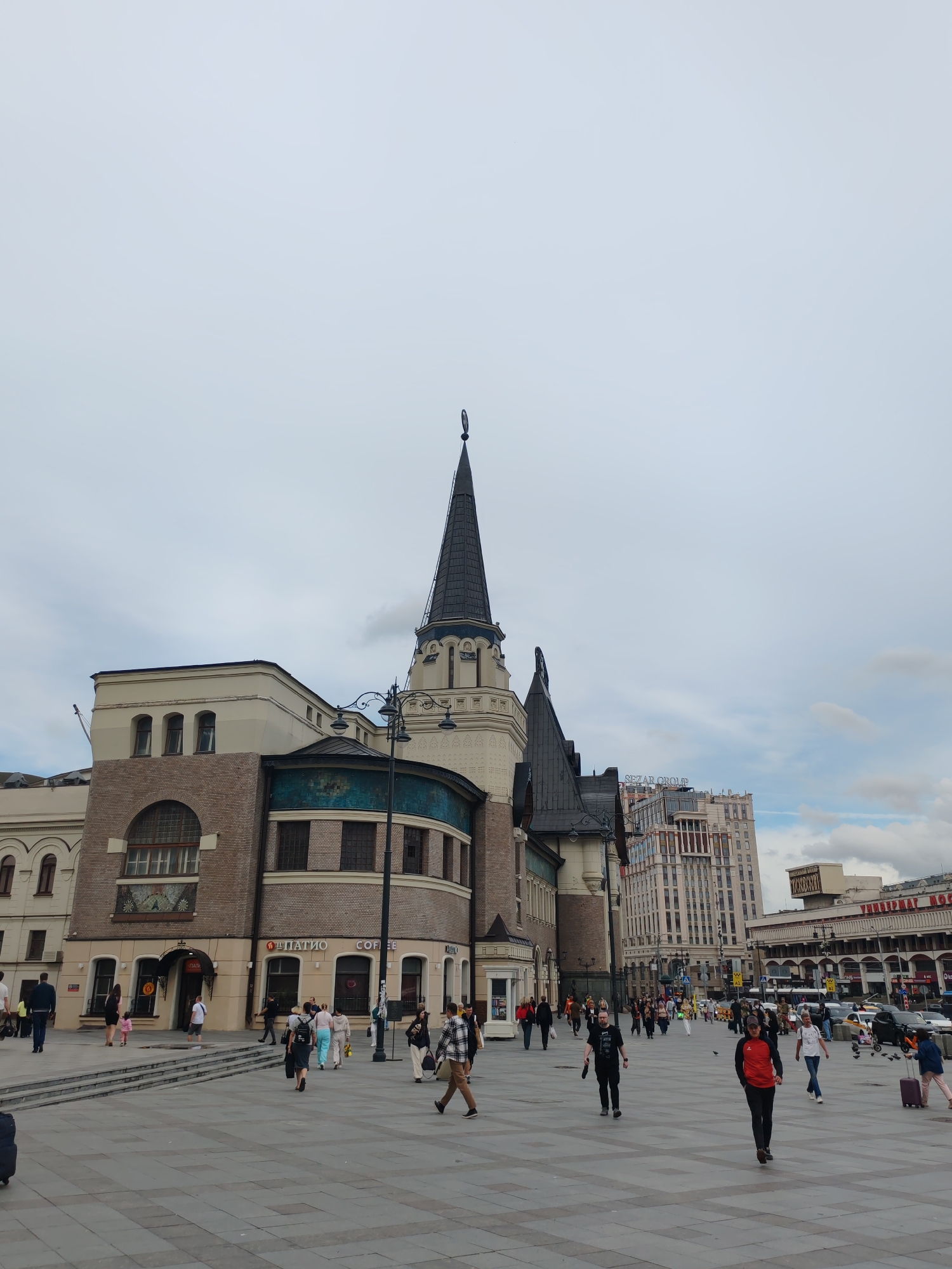 The image depicts a wide, paved plaza in front of a large building with a distinctive pointed spire, situated in Moscow, Russia. The building's architecture is a mix of old and new, with a brick facade, arched windows, and a cream-colored upper section. Several people are walking across the plaza, some with luggage, indicating it could be a transportation hub. Behind the spire building, taller, modern office buildings are visible, one with the "SEZAR GROUP" logo. The sky is overcast, suggesting a cloudy day. The plaza is well-maintained with a patterned pavement and streetlights. The scene appears to be a public space bustling with pedestrian activity.