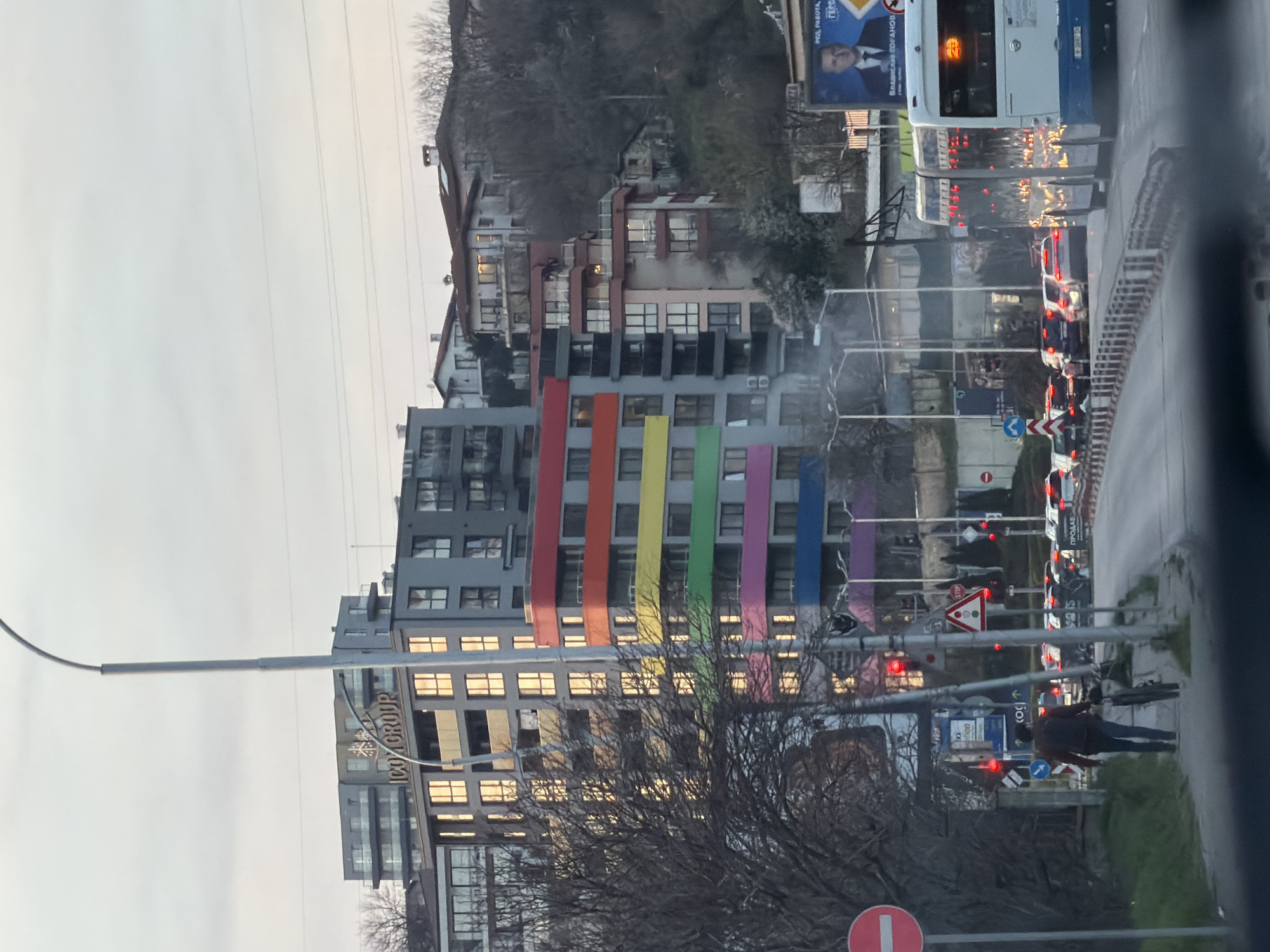 The image shows an urban street scene in Varna, Bulgaria. A modern, multi-story building with a gray facade is prominently visible, distinguished by vertical stripes in rainbow colors (red, orange, yellow, green, blue, purple). The building appears to house commercial space as the words "ICOM GROUP" are displayed on its side. A street light is in front of the building on the left side of the frame.
In the background, there is a hill covered with residential buildings and sparse vegetation. A white and blue bus is stopped in traffic. Several cars are lined up behind the bus, with their brake lights illuminating the street. On the sidewalk, a person wearing a dark jacket is walking from left to right. Traffic signs and other street furniture are also visible. The weather appears overcast.