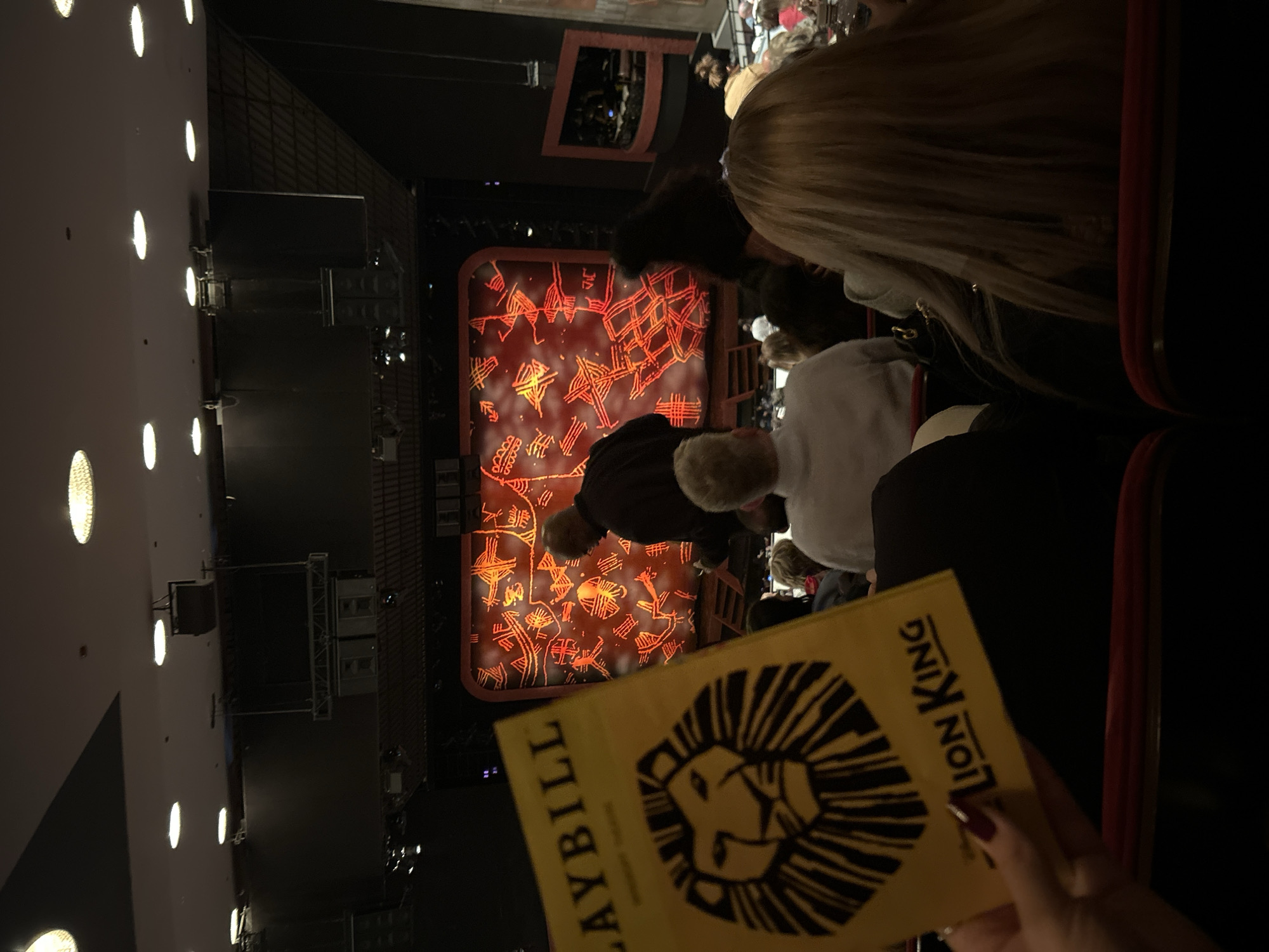 The image shows an audience view inside a theater in New York City, United States of America, presumably during a performance of "The Lion King." A Playbill for the show is prominently displayed. The stage is visible in the background with orange and red patterns. Seated audience members are also visible, including some with fair skin and blond hair. Overhead lighting is visible on the left side of the image.