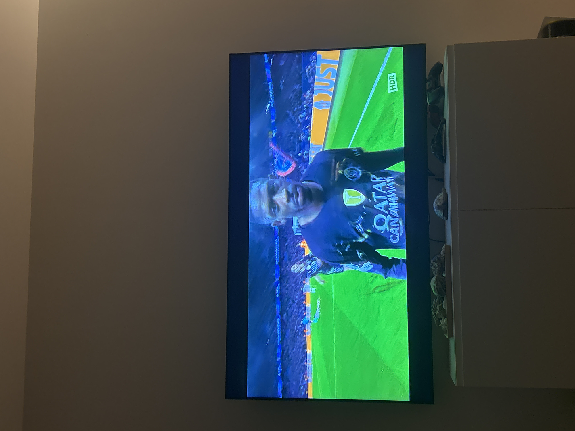 A television displays a soccer game, featuring a player in a blue uniform with yellow accents and the word "QATAR" visible. The game is set on a green field. The TV is mounted on a wall next to a white cabinet. The location in Villefranche-sur-Mer, France is not evident from the image.