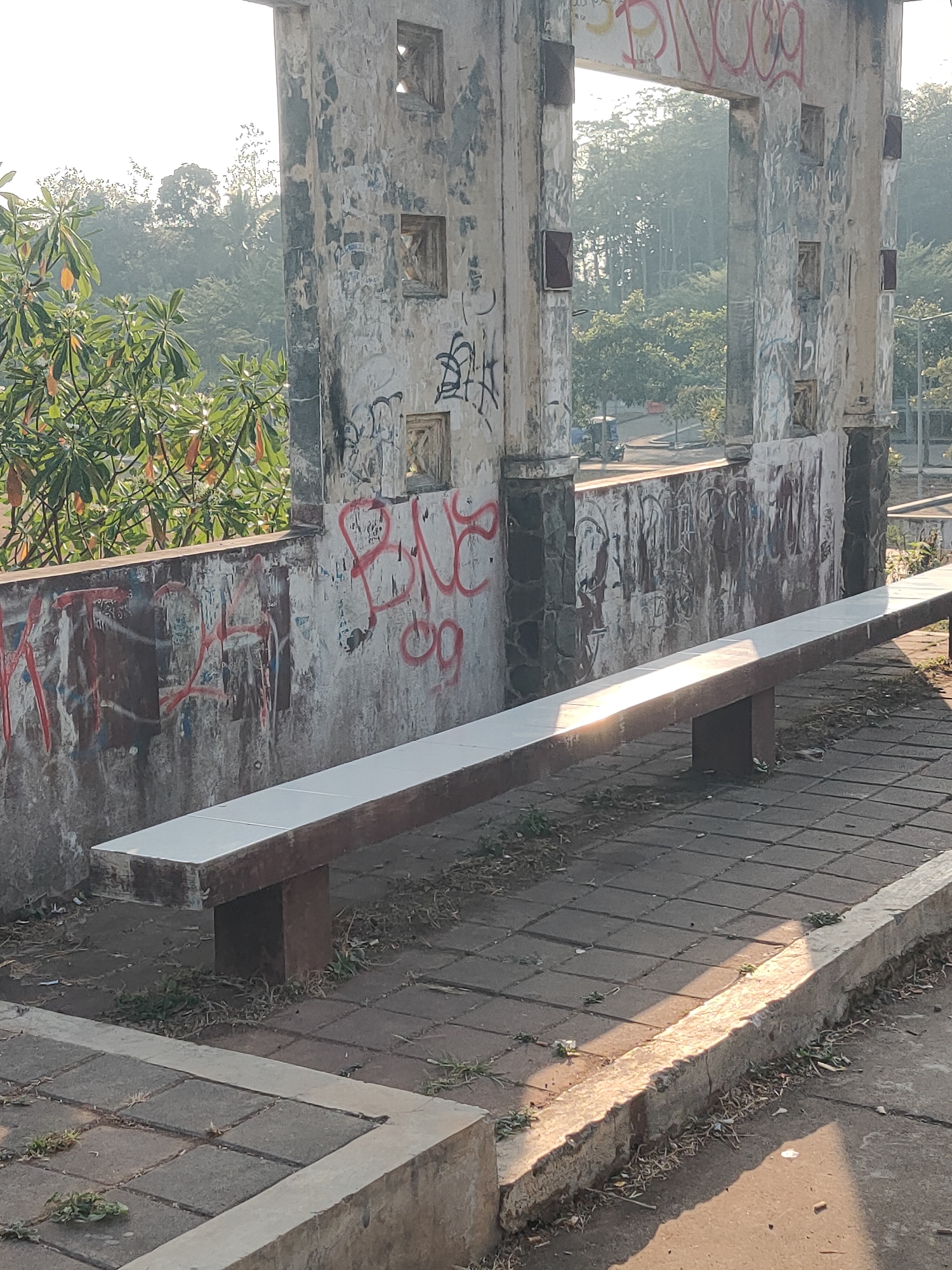 The image depicts a sunlit outdoor scene featuring a long, white-tiled bench with dark brown supports, positioned on a patterned grey paver pathway. The bench is set against a weathered, graffiti-covered wall constructed of concrete and stone. Behind the wall, lush green foliage and trees are visible, suggesting a natural or park-like environment. The overall aesthetic is one of urban decay and nature's reclamation, with graffiti on the wall and grass growing through the pavers. The lighting indicates it is daytime, with sunlight casting strong shadows across the scene. The image does not contain any people or actions.