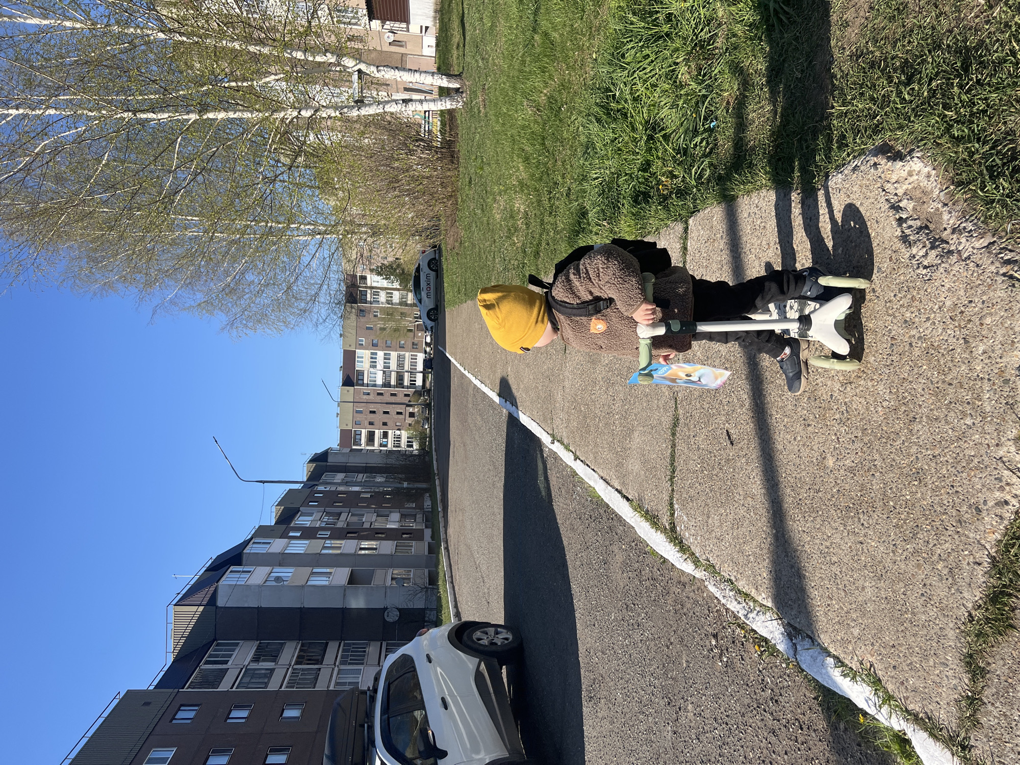 A child in a yellow hat and brown coat is on a scooter on a paved path with a white painted curb. Behind them are apartment buildings, trees, and parked cars. The location is Марковский, Russia.
