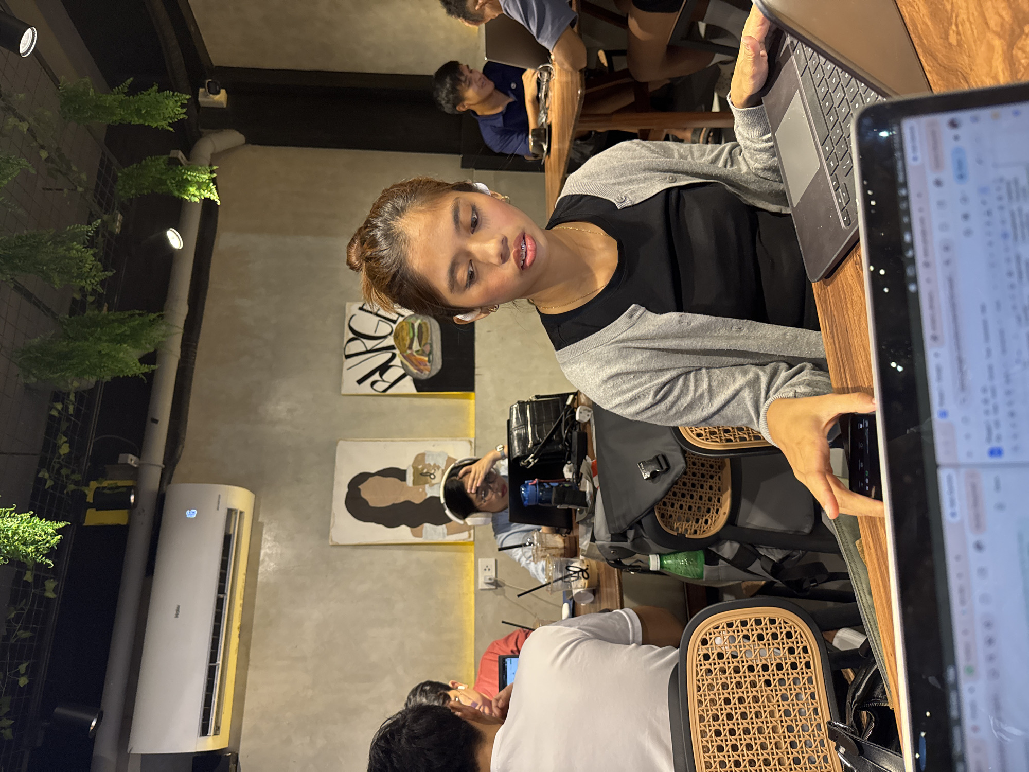 The image depicts the interior of a café in Davao City, Philippines. A young woman with brown hair in a bun is working on a tablet and a laptop at a wooden table. She is wearing a grey cardigan over a black top and earbuds. Several other people are present in the café: other patrons are seated at nearby tables, working on their laptops or conversing. The café has white walls with decorative paintings, track lighting, and greenery hanging from the ceiling. An air conditioning unit is mounted on the wall.