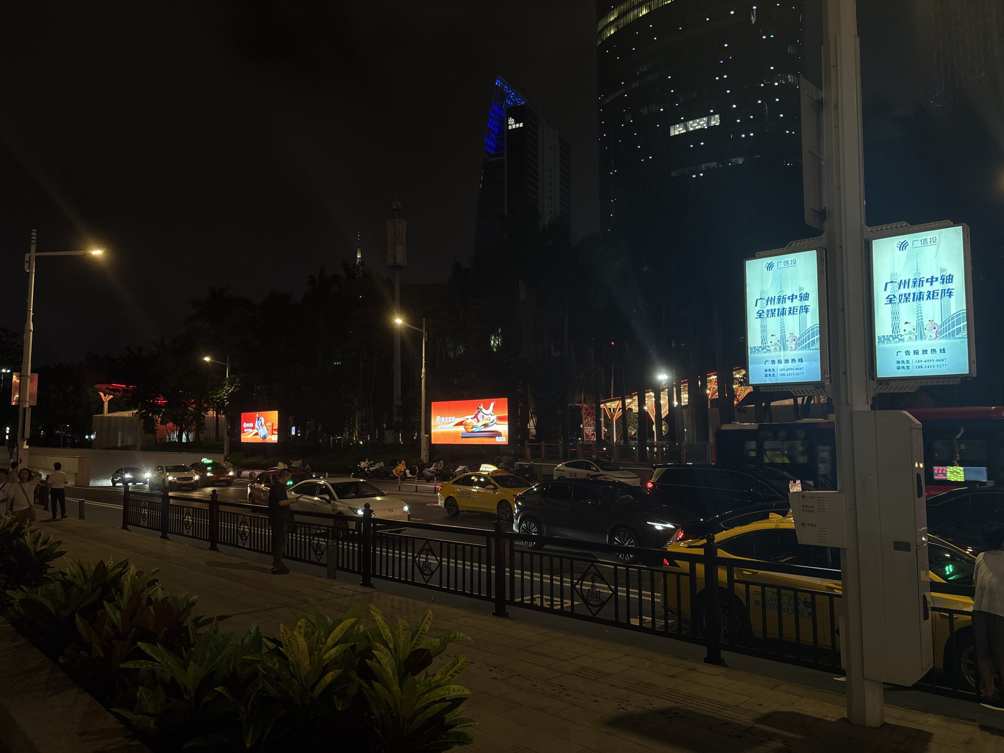 This media file captures a nighttime scene on a city street, likely in Tianhe District, China. The foreground features lush, dark green foliage, hinting at a warm and possibly humid climate. Beyond the foliage, a sidewalk with a black metal railing separates pedestrians from a busy road. Several cars, including yellow taxis and a dark SUV, are visible on the road, suggesting a lively urban environment.
Towering buildings dominate the background, with one skyscraper illuminated by blue lights, creating a striking visual contrast against the dark sky. Other buildings are dotted with numerous small lights, indicating they are occupied. Two large digital billboards are prominently displayed on a pole, one on the left and one on the right. Both billboards feature Chinese text, with the phrase "广州新中轴 全媒体矩阵" (Guangzhou New Central Axis, All Media Matrix) being the most prominent. Contact information for advertising is also visible.
Streetlights cast a warm glow on the scene, illuminating the road and sidewalk. The overall atmosphere is one of a bustling city at night, with a mix of modern architecture and everyday urban life. The presence of people on the sidewalk, some looking at their phones or walking, adds to the sense of activity. The time of day is clearly night due to the darkness and the artificial lighting. The weather appears to be clear, as there are no visible signs of rain or fog.