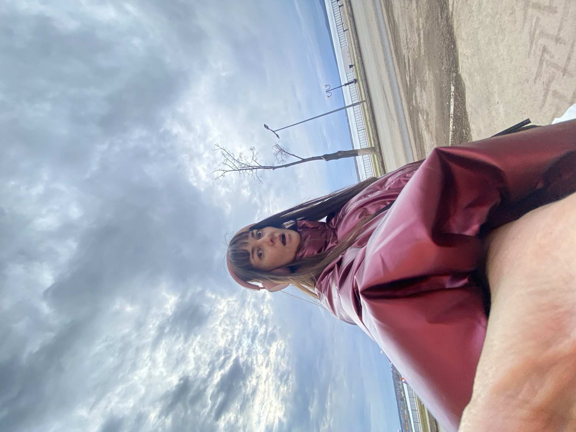 A low-angle, selfie-style shot depicts a woman wearing a shiny, reddish-purple jacket and light pink headphones. She has light skin, brown hair with bangs, and an open-mouthed expression, suggesting surprise.
The background reveals a cloudy sky above a street and a body of water. In the distance, lampposts and a barren tree stand along the street. The street and water suggest an urban location. According to context clues, the location is the city of Votkinsk, Russia.
