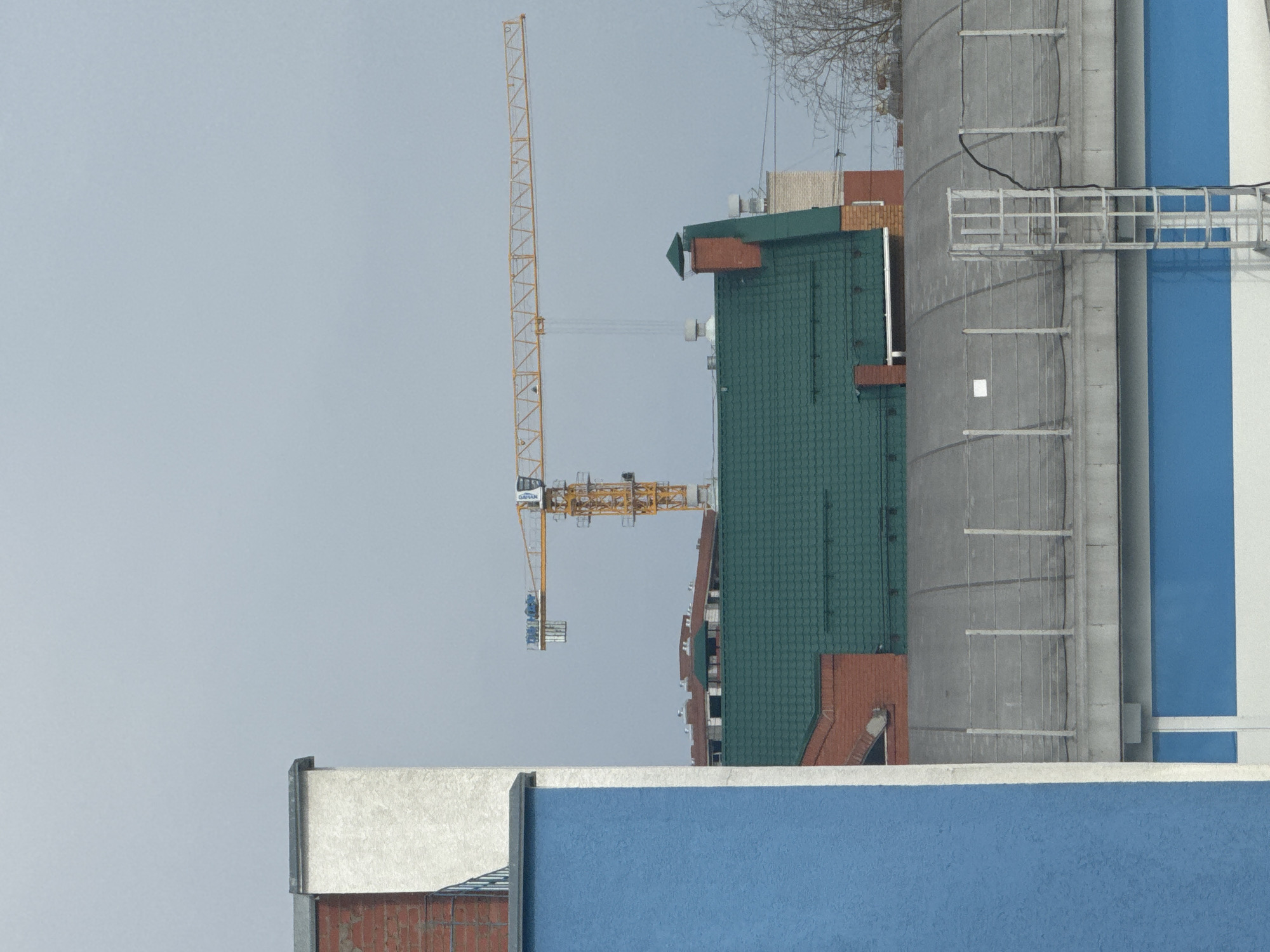 The image depicts a construction scene in the city of Chaikovsky, Russia. A yellow tower crane is positioned adjacent to a building with a green tile roof and red brick accents. To the right of the green roof building is a taller concrete structure with a blue and white vertical stripe, featuring external ladders and a platform. In the foreground, a blue and white wall partially obscures the view of the buildings. The sky is overcast, suggesting a cloudy or hazy day. There is no visible presence of people.