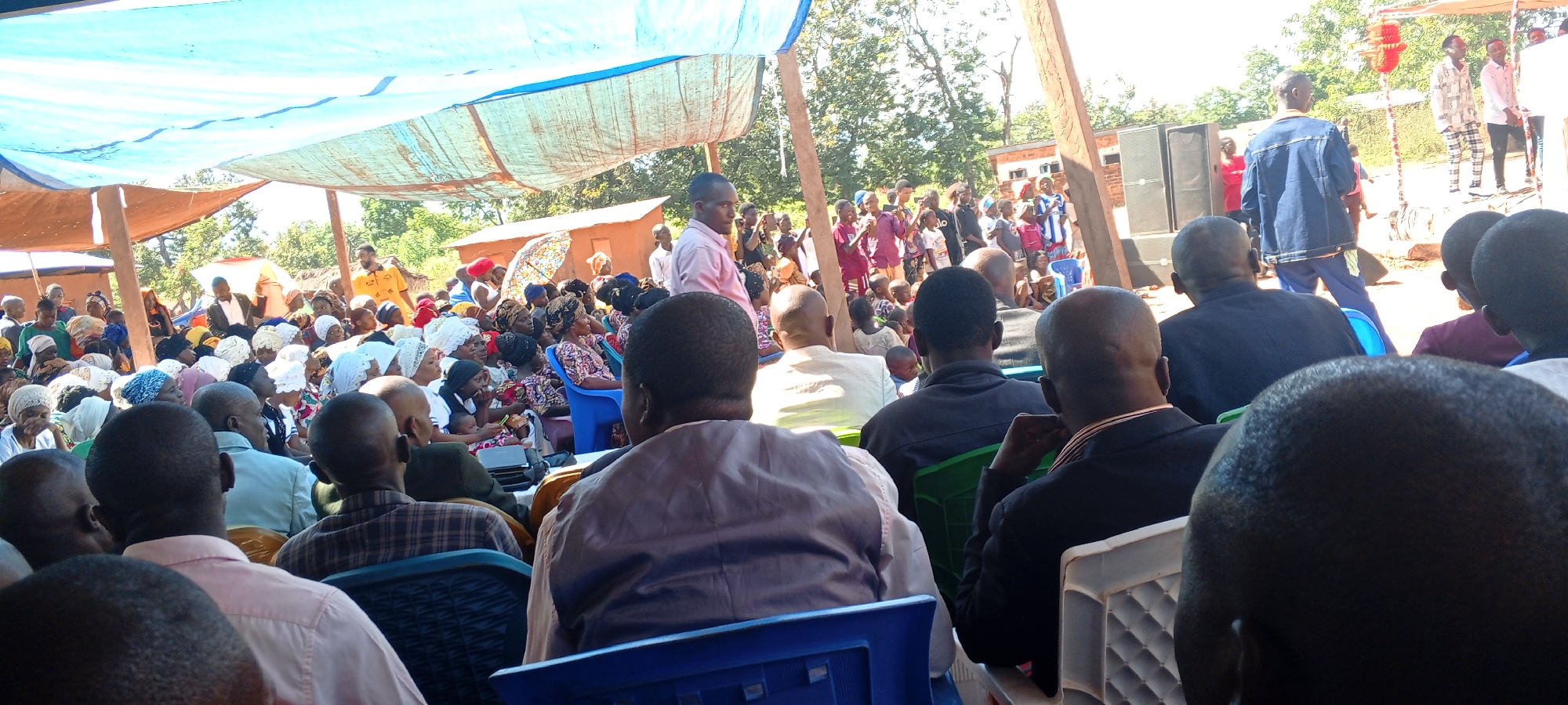 The image shows a large gathering of people, mostly sitting, under a canopy in an outdoor setting. Many attendees are African. Some individuals are standing on a raised platform at the front, and there are speakers flanking the platform. The environment appears to be rural, with trees and simple buildings visible in the background. This scene is likely taking place in the city of Kalemie, Democratic Republic of the Congo.